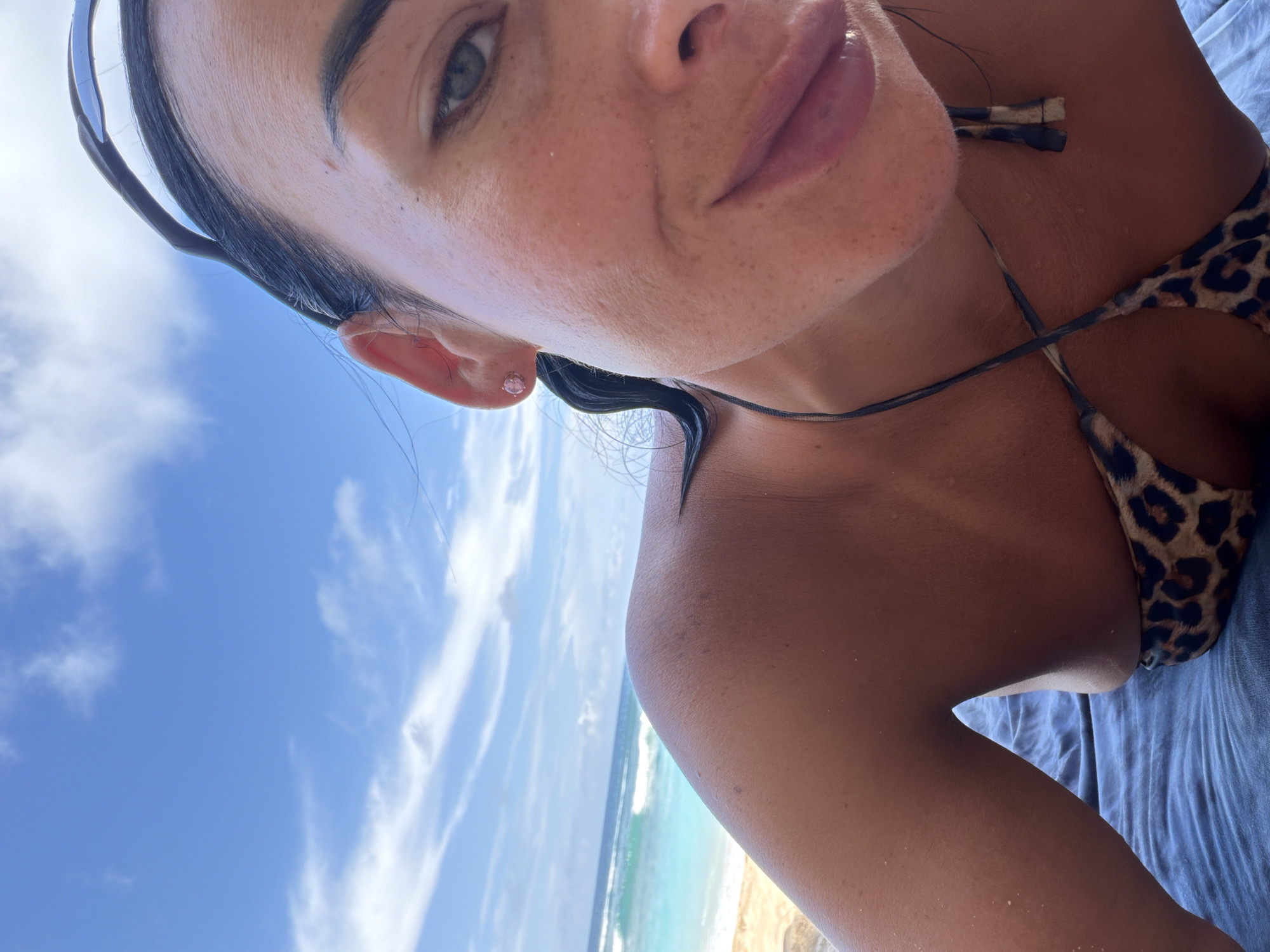 A tanned woman with dark hair and freckles is lying down, likely on a beach. She is wearing a leopard-print bikini top and has sunglasses pushed up on her forehead. The background reveals a tropical scene with a sandy beach, turquoise water, and a blue sky with some clouds. The image suggests the scene is likely in a coastal area, possibly Ungasan, Indonesia.