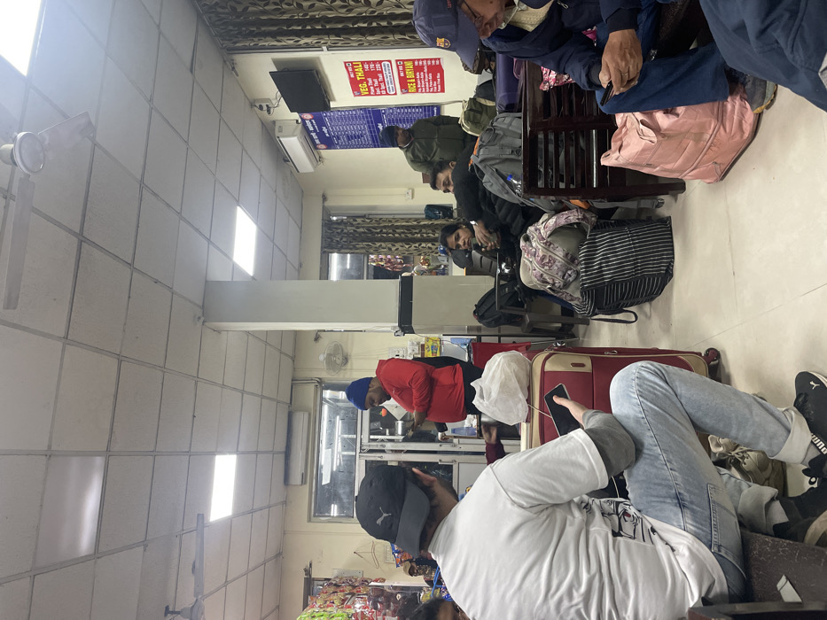 The image captures an indoor waiting area, likely a small restaurant or a waiting hall at a transit hub in Haldwani, India. The setting is functional, featuring light-colored, tiled walls and a ceiling with multiple rectangular fluorescent lights providing illumination.
Several individuals, mostly men, are present in the room. They are dressed in warm, casual clothing such as jackets, hoodies, and caps, suggesting cool or cold weather. Most are seated on simple wooden benches or chairs, appearing to be resting or patiently waiting. Some individuals are engaged with their mobile phones. Various pieces of luggage, including suitcases, duffel bags, and backpacks, are scattered near the seated people, indicating travel. One person wearing a red jacket and a blue turban is standing near what appears to be a counter area.
Visible objects include ceiling fans, a wall-mounted air conditioner unit, and a small television screen mounted high on a wall. A prominent blue menu board displays food items in both Hindi and English. Clearly legible text includes "VEG THALI" with prices such as "170/-", "150/-", and "140/-", as well as "RICE & BIRYANI" with prices like "170/-", "150/-", and "130/-". Other partially visible Hindi text reads "सूप और ..." (Soup and ...). The artificial lighting suggests the time of day could be evening or night, or simply an indoor setting where natural light is limited. The overall impression is a common waiting area or eatery catering to travelers.