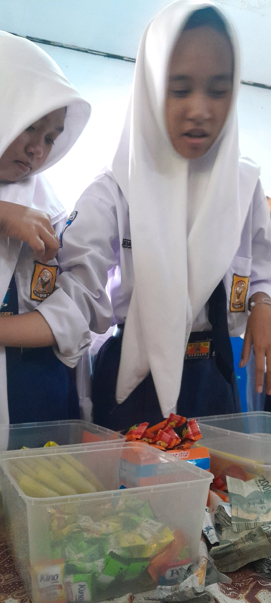 In Tahunan, Indonesia, two girls wearing white hijabs and school uniforms are partially visible. The girl on the left is adjusting her uniform patch, while the girl on the right looks down. In front of them are two clear plastic containers filled with various items. The container on the left holds what appear to be snacks and wrappers, including some yellow cylindrical items. The container on the right contains more colorful snack wrappers, predominantly red and orange, and some loose candies. To the right of the containers, a stack of Indonesian Rupiah banknotes is visible. The scene appears to be indoors, with a light-colored ceiling in the background.