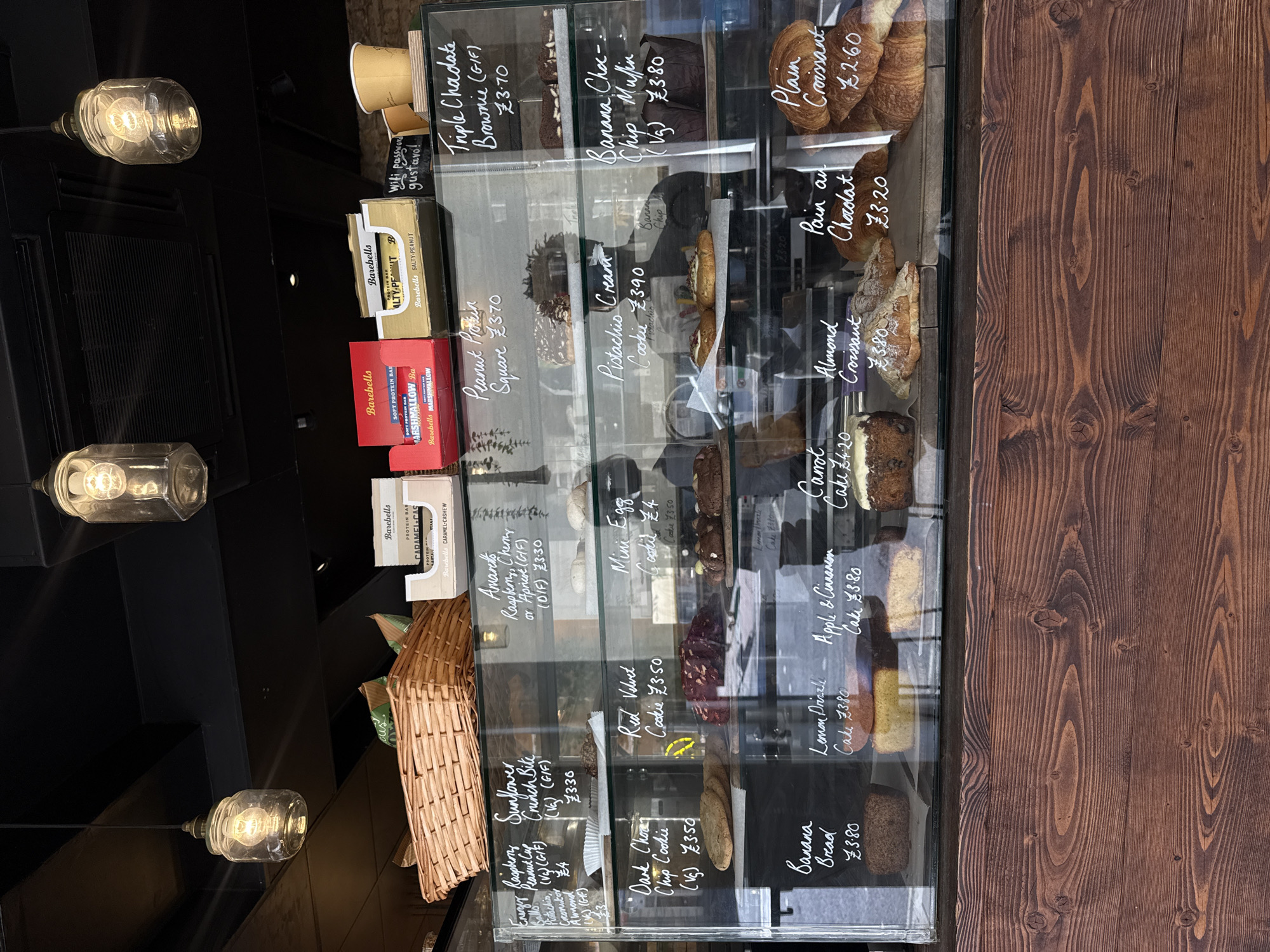 The image shows a bakery display case in London, United Kingdom. The case contains various pastries and baked goods, including brownies, muffins, cookies, croissants, and cakes. Prices are listed for each item. The display case is against a dark wooden wall. There are some protein bars, boxes, and a basket with some more products near the bottom left. There are also some light fixtures hanging from the ceiling. The pastry products are mostly visible on the right-hand side of the display case.