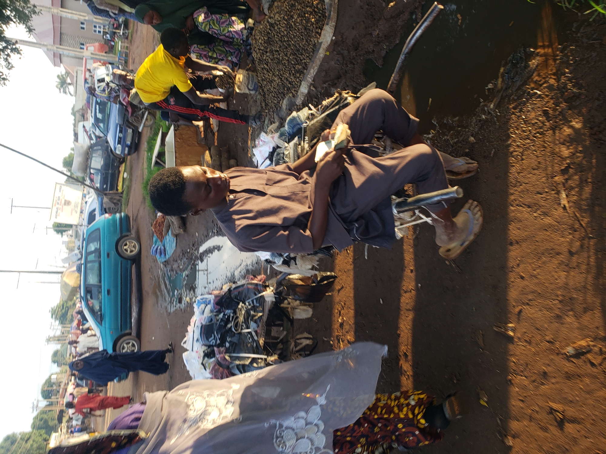 The image depicts an outdoor market scene in Kakuri Gwari, Nigeria, with a focus on a young man sitting on a stool and holding cash. The man, dressed in a brown tunic and trousers, appears to be a vendor or customer, counting money in his hands. He is situated amidst piles of goods, possibly clothing and other merchandise, some of which are bundled in plastic bags. To his right, a large mound of small, brown items, likely nuts or seeds, is visible. In the background, several vehicles are parked on a dirt road, including a blue station wagon and a black SUV. Other individuals, dressed in traditional attire, are dispersed throughout the market, some walking and others engaged in activities. The ground is predominantly dirt, with puddles of water indicating recent rain or a water source. The overall atmosphere suggests a busy, informal marketplace under natural lighting conditions, possibly late afternoon given the shadows.