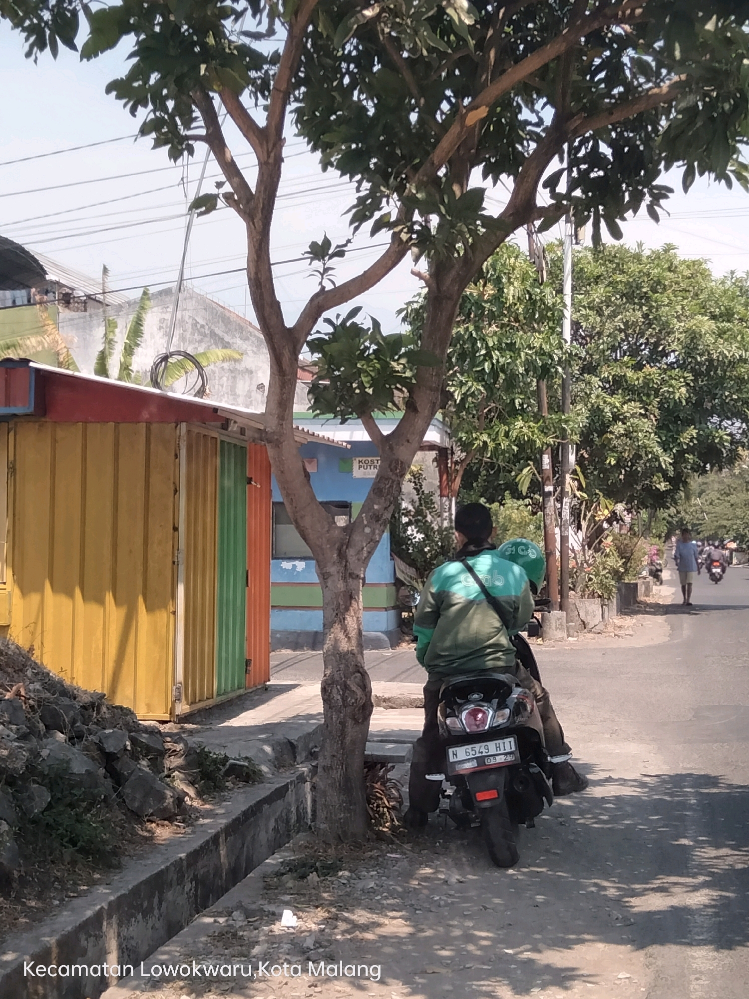 The image depicts a street scene in the Lowokwaru District of Malang, Indonesia. In the foreground, a person is seated on a motorcycle, wearing a green jacket with the "grab" logo. The motorcycle's license plate reads "N 6549 HII" with an expiration date of "09-29". To the left, there is a building with colorful corrugated metal walls in yellow, green, and orange, and a red roof trim. A tree with sparse foliage stands between the motorcycle and the building. A concrete drainage ditch runs along the side of the road. In the background, more trees and buildings are visible, and other motorcycles and pedestrians are present on the road, suggesting a moderately busy urban environment. The sky is clear and blue, indicating fair weather.