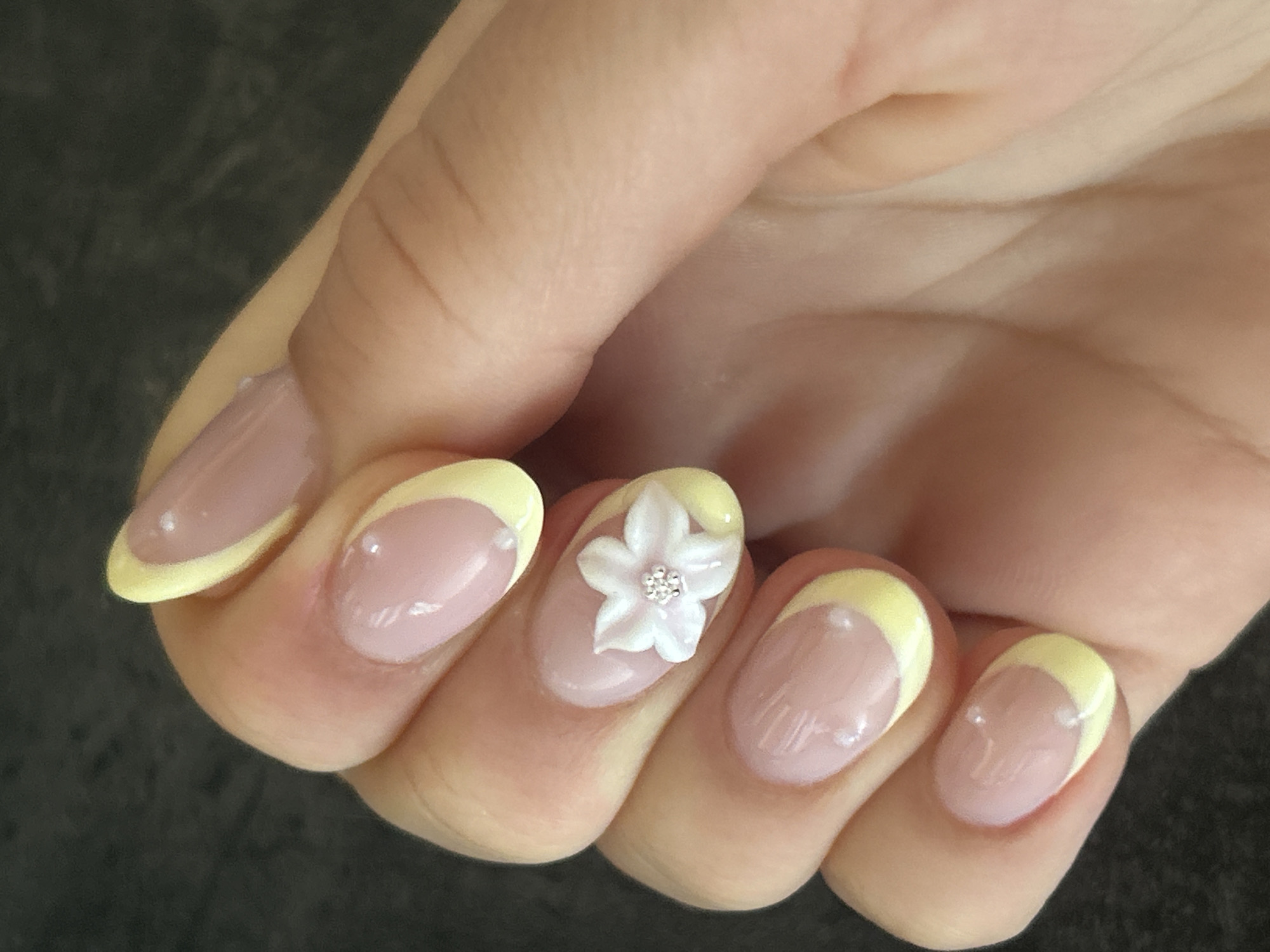 A close-up, top-down view of a person's left hand displays a French manicure with a pale yellow tip. The nails are oval-shaped and have a sheer pink base coat. The middle finger on the right side of the image features a small, white, three-dimensional flower with a cluster of tiny silver rhinestones in its center. The background is a dark, textured surface, possibly a table or countertop. The lighting is bright, creating subtle reflections on the nail polish.