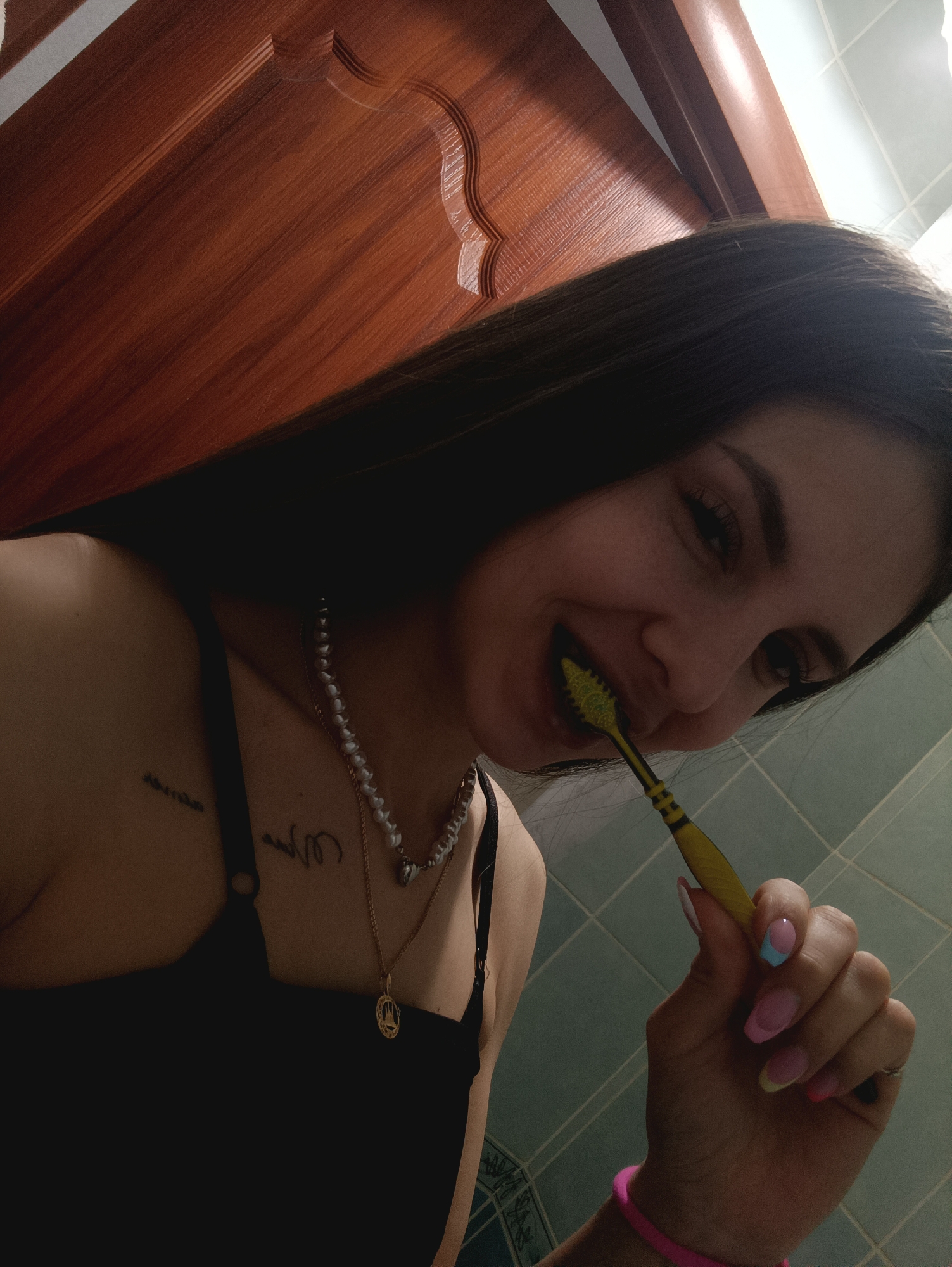 A young woman is brushing her teeth in a bathroom with light-blue tiled walls and a wooden cabinet. She is wearing a black top and several necklaces. Her fingernails are painted with pastel colors. She is holding a yellow toothbrush in her right hand. The location is the city of Unknown, Unknown.