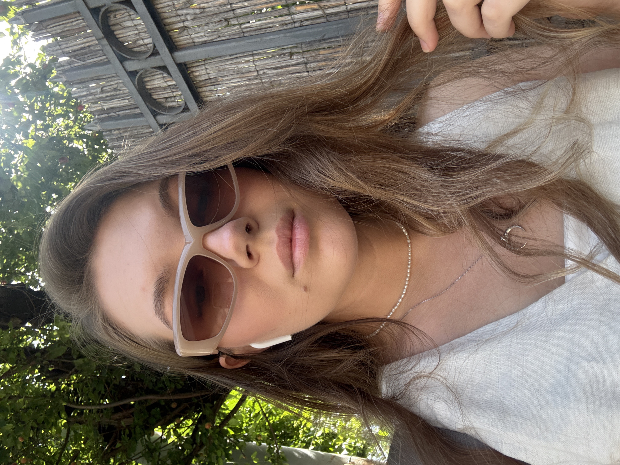 A young woman with long, brown hair and fair skin is captured in a close-up, selfie-style photograph. She is wearing large, beige-framed sunglasses with brown lenses, and white wireless earbuds. A delicate pearl necklace and a silver chain with a crescent moon pendant are visible around her neck. She is dressed in a white linen top. Her right hand is casually touching her hair. The background features lush green foliage and a rustic wooden fence with metal accents, suggesting an outdoor urban setting. The sunlight filters through the leaves, creating a bright and airy atmosphere. The location is identified as Vozdovac Urban Municipality, Serbia.