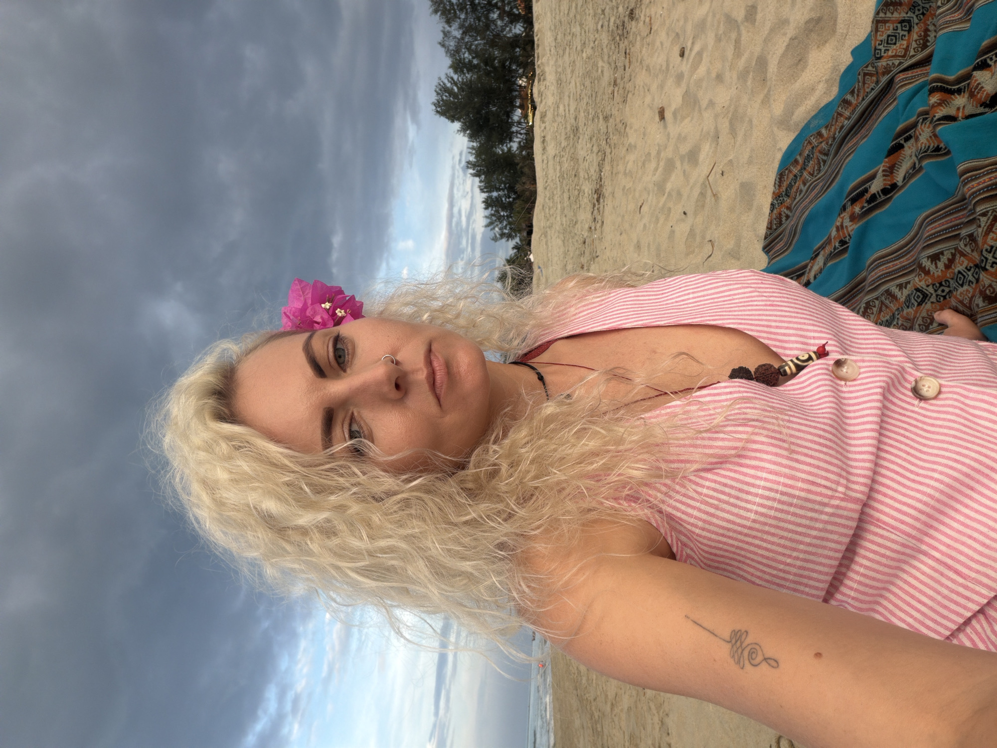 A woman with blonde, curly hair adorned with a pink flower sits on a sandy beach, facing forward. She wears a pink and white striped sleeveless dress with buttons down the front. A tattoo is visible on her left arm, and she has a nose ring. Behind her, the ocean stretches to the horizon under a cloudy sky. Trees line the background, and a patterned blanket is spread on the sand beside her. This scene is located at An Bang Beach in Hoi An, Vietnam.