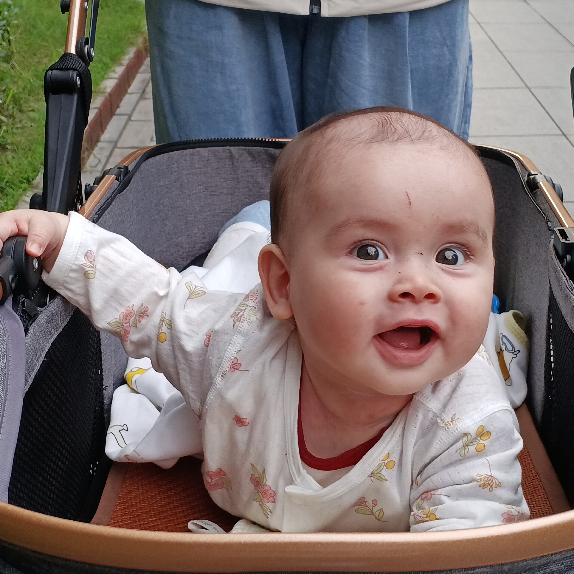 An overhead shot captures a joyful baby lying on its stomach in a stroller, looking directly forward with wide, curious eyes and an open-mouthed laugh or smile. The baby, with light skin and dark hair, has a small red scratch on its forehead. It is dressed in a white long-sleeved onesie adorned with pink and yellow floral patterns. White socks with a yellow and white cartoon animal design are also visible on its feet.
The stroller features a grey fabric body with brown/bronze frame elements and black accents. A mesh pocket is visible on the side. The baby's left hand is gripping a black part of the stroller's frame.
Partially visible in the background is the lower half of an adult, likely pushing the stroller, wearing blue pants. The setting appears to be outdoors on a paved pathway, with green grass visible to the left. The scene suggests a pleasant daytime stroll in an urban or suburban area of Shunde District, China. There is no clearly legible text visible in the image.