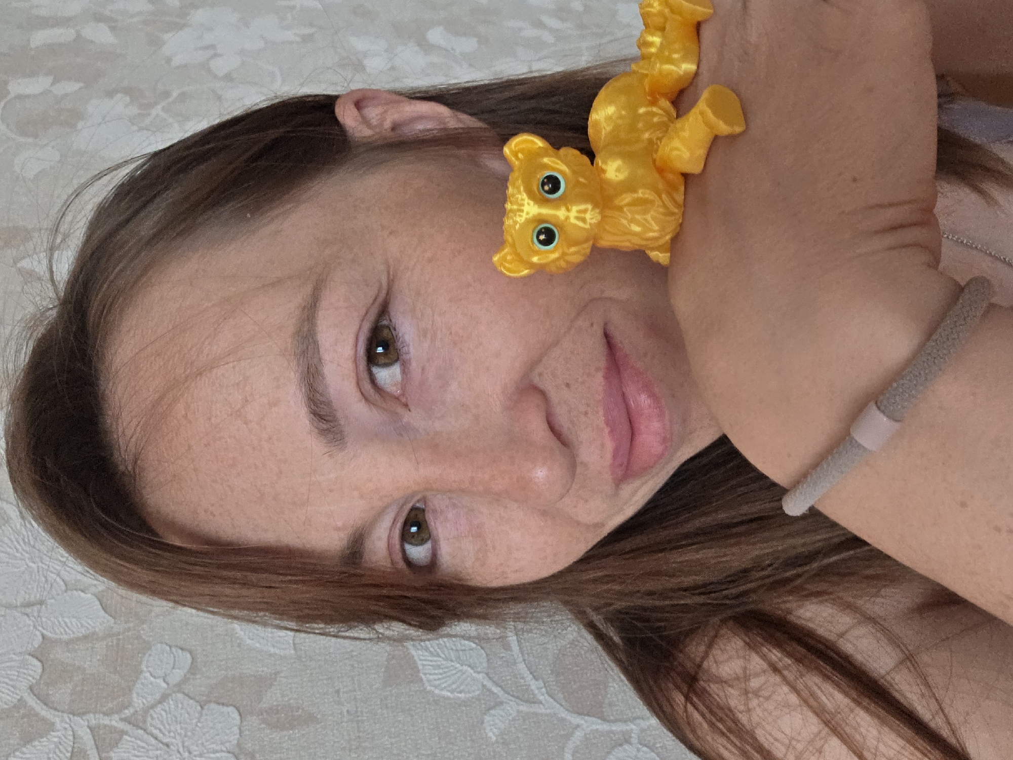 A portrait of a fair-skinned woman with brown hair and brown eyes is presented. A yellow toy cat, with large blue eyes, is attached to her forearm. She is wearing a gray bracelet on her wrist. The background features a light-colored floral patterned fabric. The photograph is taken from an angle, with the woman's face tilted. The location and city are not discernible from the image.