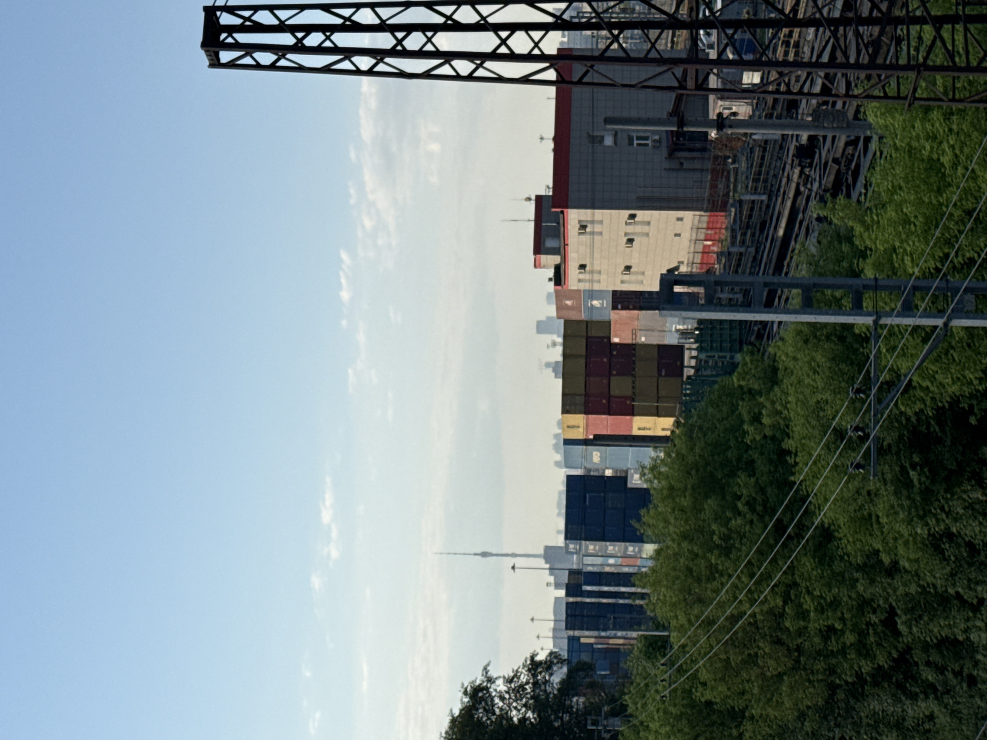 The image depicts a cityscape in Moscow, Russia. In the foreground, power lines and green foliage are visible. Behind them, stacks of colorful shipping containers create a distinct visual element. Further back, a light-colored building with a red-trimmed roof can be seen. The Ostankino Tower rises in the distance, a prominent landmark. A metal structure, possibly part of a bridge or an industrial crane, appears in the upper left corner of the frame against a light blue sky with scattered clouds.