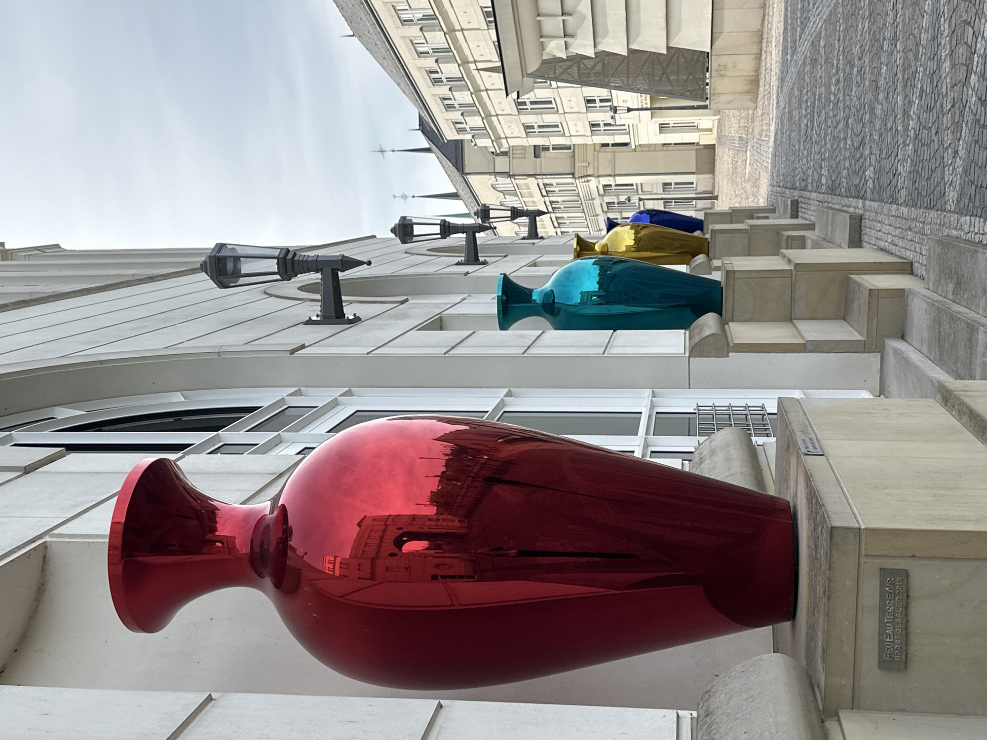 The image shows a row of large, colorful, reflective vases displayed in front of a modern building with arched windows. The largest vase, on the left, is bright red and reflects a building with an archway. To its right are smaller vases in turquoise, gold, and blue. The building has white walls and decorative lanterns mounted on the exterior. In the background, there are other buildings with classical architecture and spires. The ground is paved with cobblestones. The scene appears to be a public outdoor space in a European city, likely a pedestrian area. There are no people visible in the image. Based on the artist's signature on the base of the red vase, which reads "FEU EAU TERRE AIR HORST BLASSER 2010", this artwork is likely a part of the "Feu Eau Terre Air" installation by Horst Blasser. This particular artwork, a series of large decorative vases, is located in Luxembourg City, Luxembourg.