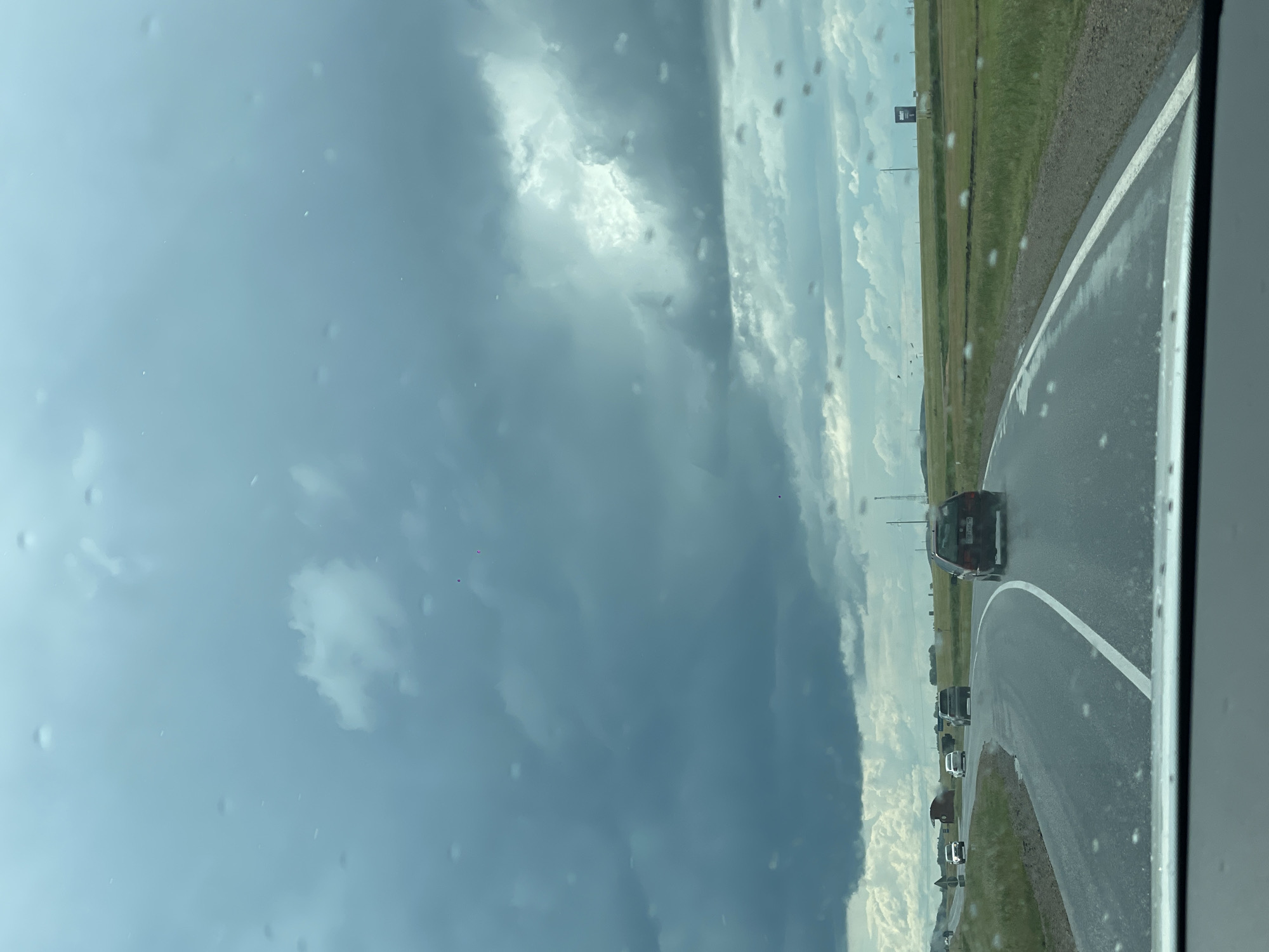 The image shows a view from inside a vehicle, looking out at a highway scene with a dark, cloudy sky. Water droplets are visible on the windshield. Several cars are driving on the road, which curves to the right. The landscape consists of green fields and distant trees, with some industrial structures and utility poles visible in the background. The sky is filled with dramatic, dark storm clouds, with patches of lighter sky peeking through. The overall impression is one of impending rain or a recent storm. The location is identified as being on the highway near the village of Zhañaaul, Kazakhstan.