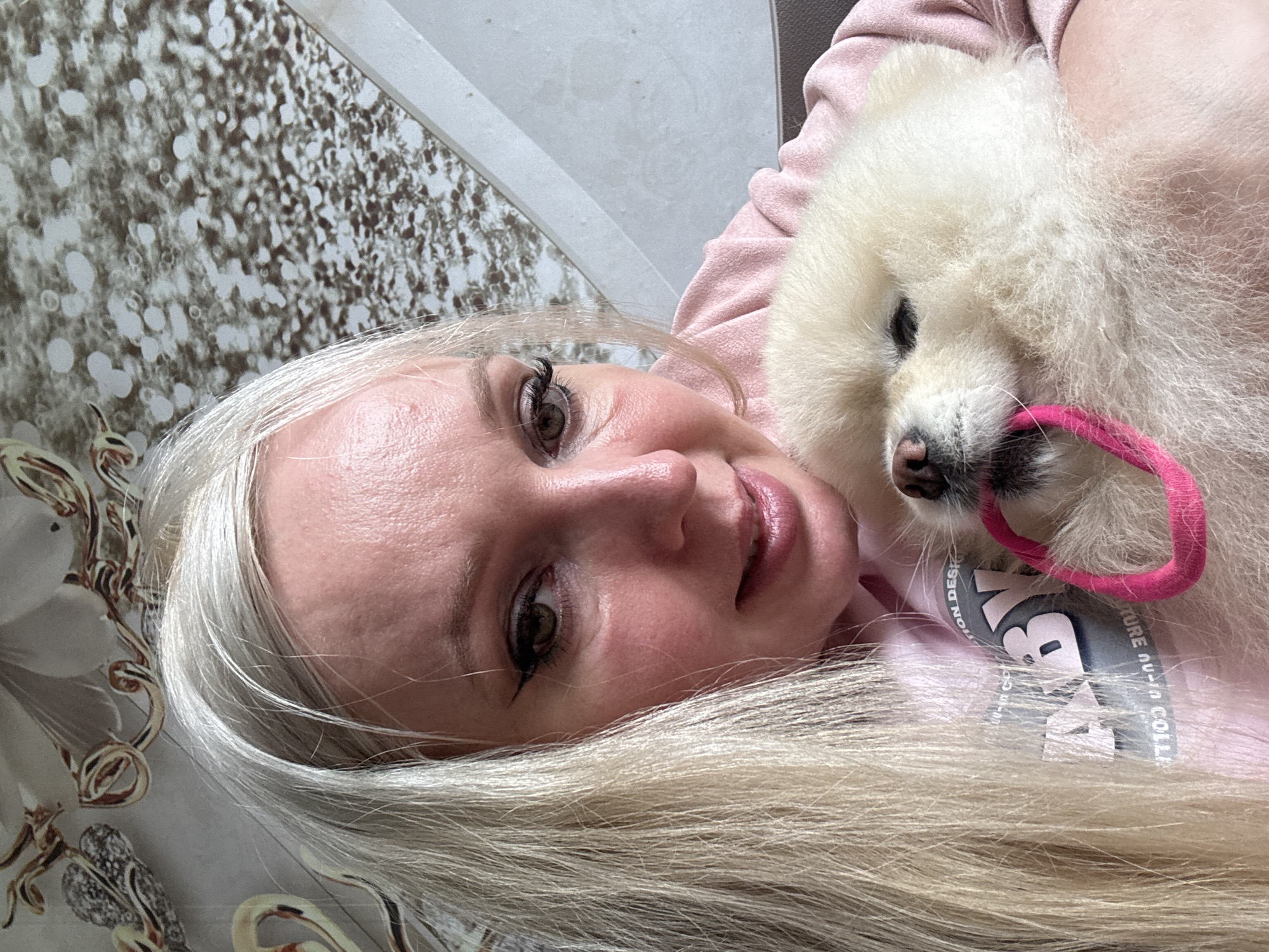 In Komsomolsk-on-Amur, Russia, a young Caucasian woman with long blonde hair and dark eyeliner is holding a fluffy cream-colored Pomeranian dog. The woman is wearing a pink hoodie. The dog is chewing on a pink ring. They are indoors, with a patterned wallpaper visible in the background.
