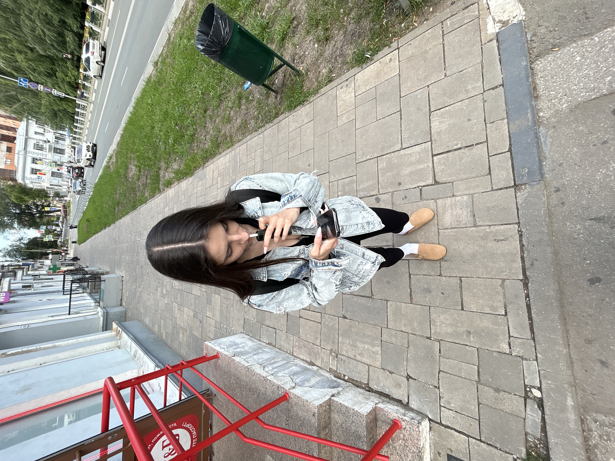 A young woman stands on a sidewalk in Novokuybyshevsk, Russia, holding a vaping device to her mouth and looking down at her smartphone. She is wearing a denim jacket, a black top, black leggings, white socks, and tan clogs. She has a backpack on. The sidewalk is paved with light gray rectangular stones. To her left is a building with a light blue facade and red railings leading up to an entrance. To her right, across a grassy strip, is a road with cars. A green trash can with a black liner is visible in the grassy area. The sky is overcast.
