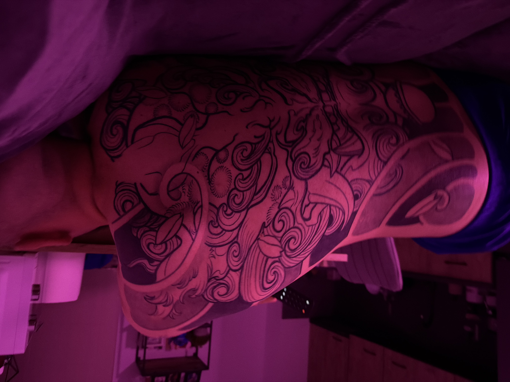 The image depicts a close-up view of a person's back and shoulder covered in an intricate black tattoo. The tattoo features swirling patterns, possibly representing water or clouds, with stylized elements that suggest a mythological or fantastical theme. The person is lying down on a surface covered with dark purple fabric, which appears to be a bed or sofa. The lighting is dim and cast in a strong magenta hue, creating a dramatic and somewhat surreal atmosphere. In the background, out of focus, are hints of a room's interior, including what appear to be kitchen cabinets and a chair with a purple cushion. There are no visible landmarks or any indication of the specific location being in Plovdiv, Bulgaria. The scene is static, with no discernible action or interaction occurring.