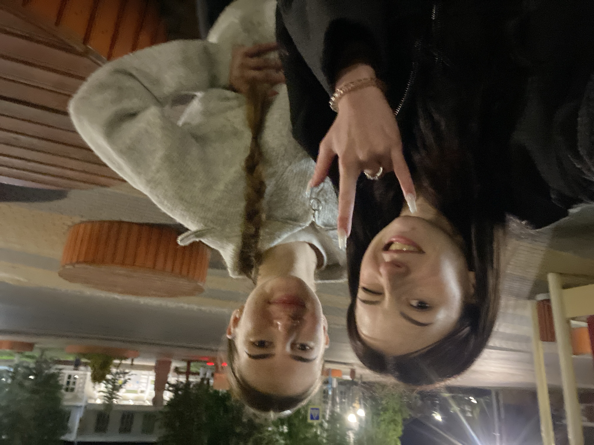 Two young women are posing for a casual photo outdoors at night. The image is oriented sideways, but if viewed upright, one woman is on the left and the other on the right. The woman on the left wears a light-colored, possibly grey, hooded garment, and has light brown hair styled in a braid that extends over her shoulder. She smiles softly. The woman on the right wears a dark jacket, has dark hair, and is winking with her right eye while showing a slight smile. Her right hand is raised, making a "rock on" or "devil horns" gesture, adorned with a ring and a beaded bracelet. Her fingernails are long and white.
The background suggests an urban or semi-urban outdoor setting at night. Elements visible include wooden slatted structures (possibly a bench or wall), a round, orange-brown object (possibly a stool or planter), and in the distance, blurry lights, trees, and buildings, indicating artificial illumination in a dark environment. The lighting suggests it is evening or nighttime, with reflections visible on the women's faces. No legible text is visible.