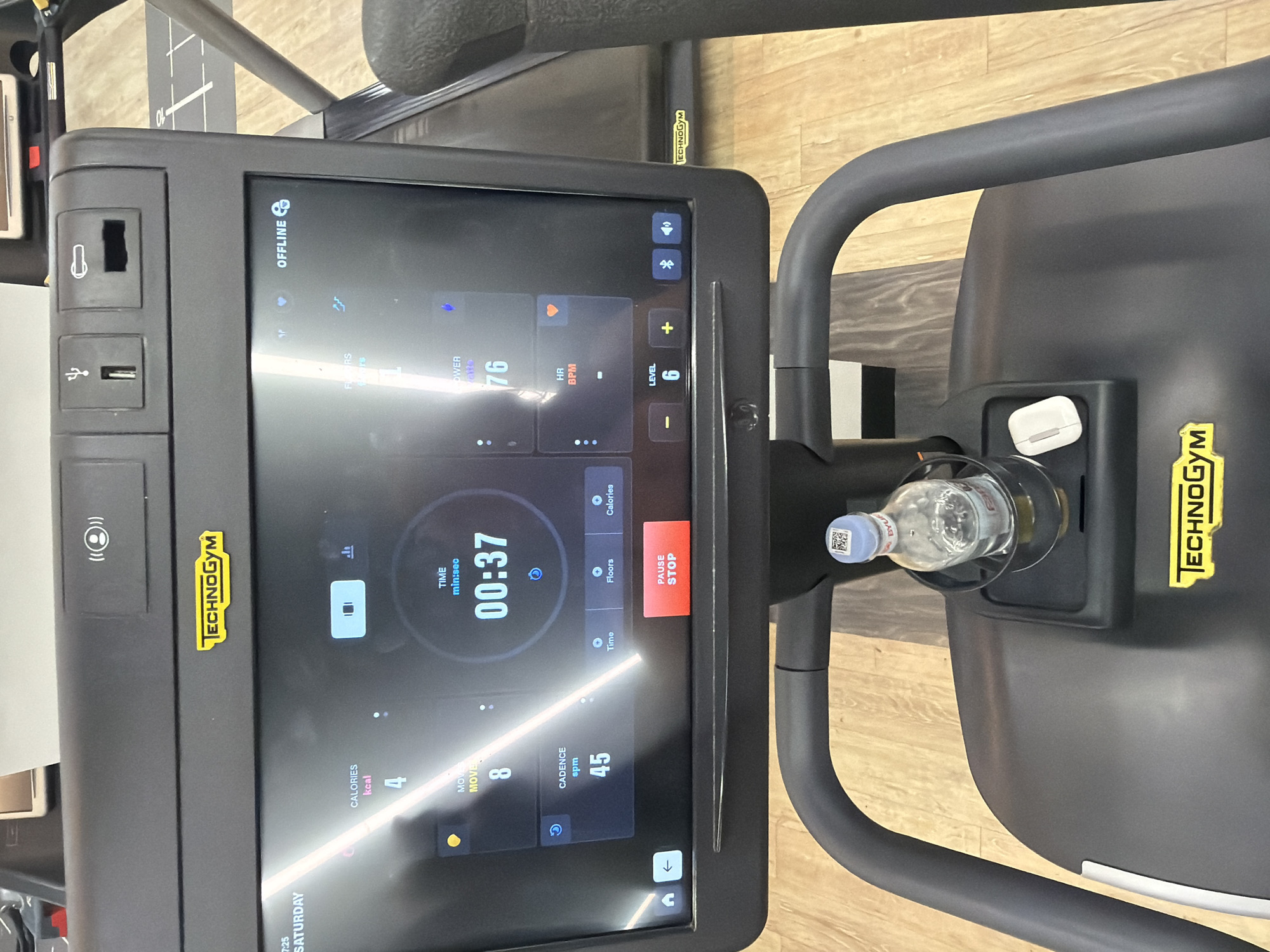The image shows the console of a Technogym treadmill. The screen displays workout information: time (00:37), calories burned (4 kcal), moves (8), cadence (45 spm), heart rate (76 BPM), and level (6). The console also features controls for pause/stop, volume, fan, and level adjustment. It displays the time as 7:25 Saturday. A water bottle is in the bottle holder, and an AirPods case is visible in a compartment. The word "OFFLINE" is displayed near the top of the console. The surroundings suggest a gym setting. The city of Yerevan, Armenia is not discernible from the image.