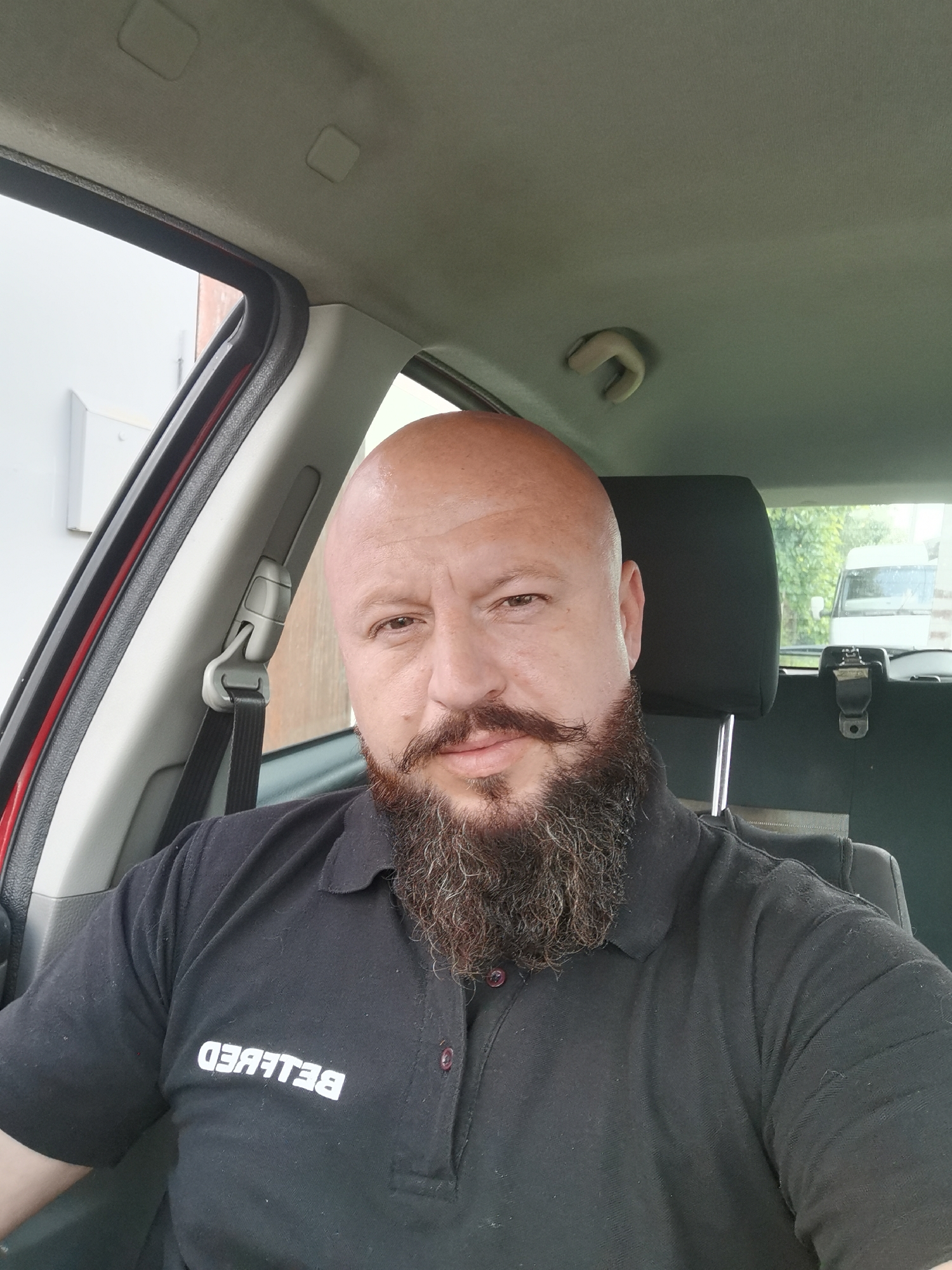 This media file is a selfie taken by a bald man with a beard and mustache, sitting in the driver's seat of a car. He is wearing a black polo shirt with the word "BELBED" visible in white text on the left side of his chest. The shirt appears to be a work uniform.
The interior of the car is visible, including the headrest of the passenger seat and the car's headliner. Through the car windows, an outdoor scene can be seen, with greenery and the back of a white van. The time of day appears to be daytime, and the weather is overcast, as indicated by the diffused light.
The man's expression is neutral, and he is looking directly at the camera. The overall impression is of a candid snapshot taken during a break or commute. The location context provided is Semypolky, Ukraine.