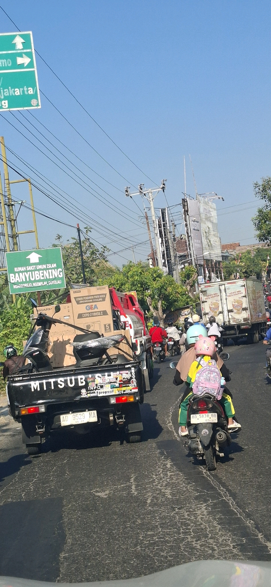 The image captures a street scene in Desa Gagaksipat, Indonesia, under a clear blue sky. A black Mitsubishi pickup truck, laden with boxes and a black scooter, is prominently positioned in the foreground, driving on an asphalt road with visible cracks. The truck's license plate reads "AD 9529 MA". To the right of the truck, a motorcycle carries two people, a woman wearing a yellow top and green pants with a pink backpack, and a child in front of her wearing a pink helmet. The motorcycle's license plate is "AD 5838 BB". Further down the road, other vehicles, including motorcycles and a large truck, are visible, indicating traffic flow. On the left side of the image, a green directional sign points to "RUMAH SAKIT UMUM ISLAM BANYUBENING". Electrical wires crisscross the sky, and power poles are scattered throughout the scene. Lush green trees and vegetation line the sides of the road. The overall atmosphere suggests a typical day with moderate traffic on a local Indonesian road.