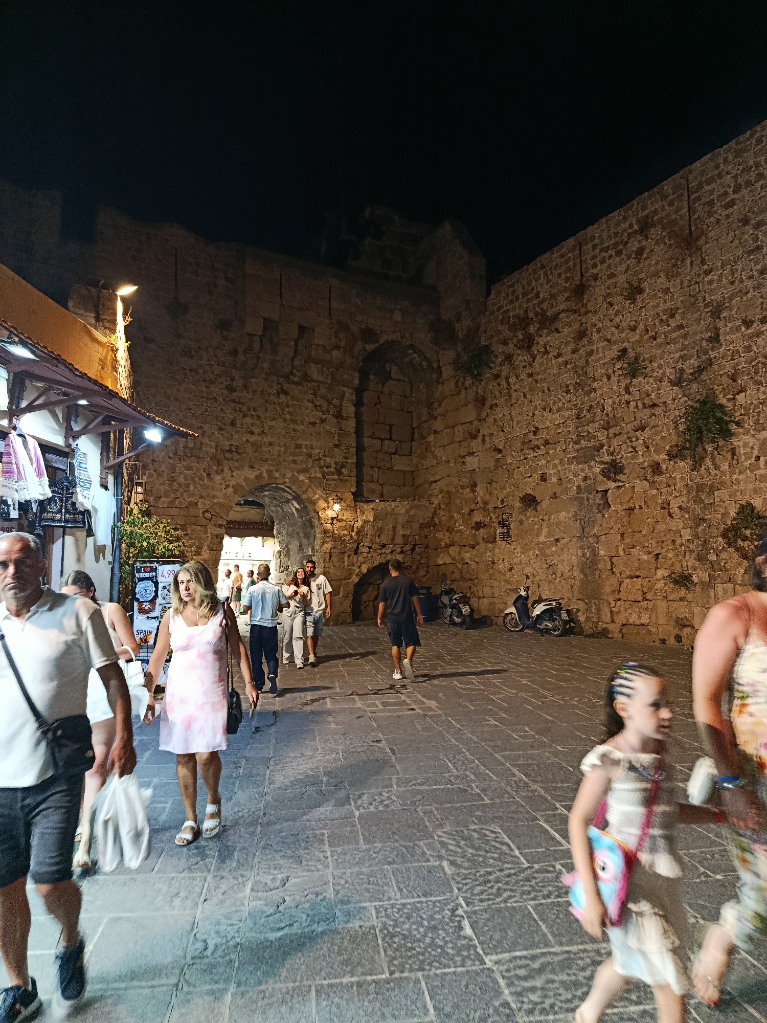 A group of people are walking along a cobblestone street at night, illuminated by artificial lights. The scene is dominated by a large, ancient stone wall, part of a fortification, with an archway in the center that serves as a passage. Shops with awnings are visible on the left side of the street, displaying various goods. Scooters are parked near the archway, suggesting a mode of transportation in the area.
The people in the foreground and mid-ground are casually dressed, some carrying shopping bags. A young girl with braided hair is walking in the right foreground, holding a colorful bag. The overall mood appears relaxed, typical of a tourist area in the evening. The stone wall is a prominent feature, indicating a historic location. The presence of shops and the attire of the people suggest a commercial and recreational area.
There is some visible text on a sign in the shop on the left, which appears to be a price list for items, with "RHODES" prominently displayed. The lighting suggests it is nighttime, and the weather appears to be clear. The architectural style of the wall and the general atmosphere strongly suggest a Mediterranean location, consistent with the provided location context of Rhodes, Greece.