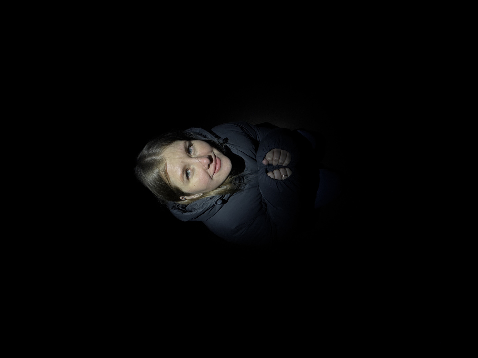 A woman with blonde hair and a grey hooded puffer jacket is centrally located in a dark environment. Her hands are clasped near her chest, and she is looking up and slightly to her left with a faint smile. The environment around her is completely black, suggesting a very dark or nighttime setting. There are no other discernible objects or individuals present. The city of Горный, Russia is not visibly identifiable in the image.