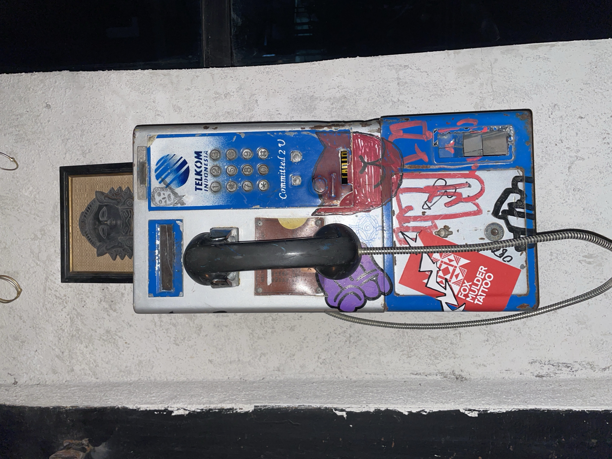 An old, weathered public payphone, likely defunct, is mounted horizontally on a light-colored, textured wall. The scene is set in what appears to be a public or semi-public space in Mengwi, Indonesia, indicated by the "TELKOM INDONESIA" branding and a traditional Balinese-style mask in a nearby framed picture.
The payphone unit is a mix of blue, white, and silver, showing significant signs of wear, rust, and colorful graffiti. Various tags and scribbles in red, black, and white cover parts of its surface. The black receiver is resting in its cradle, connected to the main unit by a coiled metal cord.
Visible text on the payphone includes the "TELKOM INDONESIA" logo and company name, as well as the slogan "Committed 2 U" displayed below the standard numeric keypad (0-9, *, #). A distinct red sticker on the right side of the unit reads "FOX MULDER TATTOO" and features a geometric pattern. A small purple cartoon hand sticker is also visible near the receiver.
To the left of the payphone, a small, dark-framed picture houses an intricate carving or drawing of a traditional Balinese mask or face. Further left, several small, round metal rings are affixed to the wall. The wall itself is rough, light-colored, and extends upwards into a dark, possibly reflective, area, suggesting a window or shadowed space. Below the wall, a thick, roughly painted black surface forms the lower part of the scene. The dim lighting suggests the image was captured at night or in an enclosed space. No specific activity is taking place, conveying a sense of urban decay or disuse.