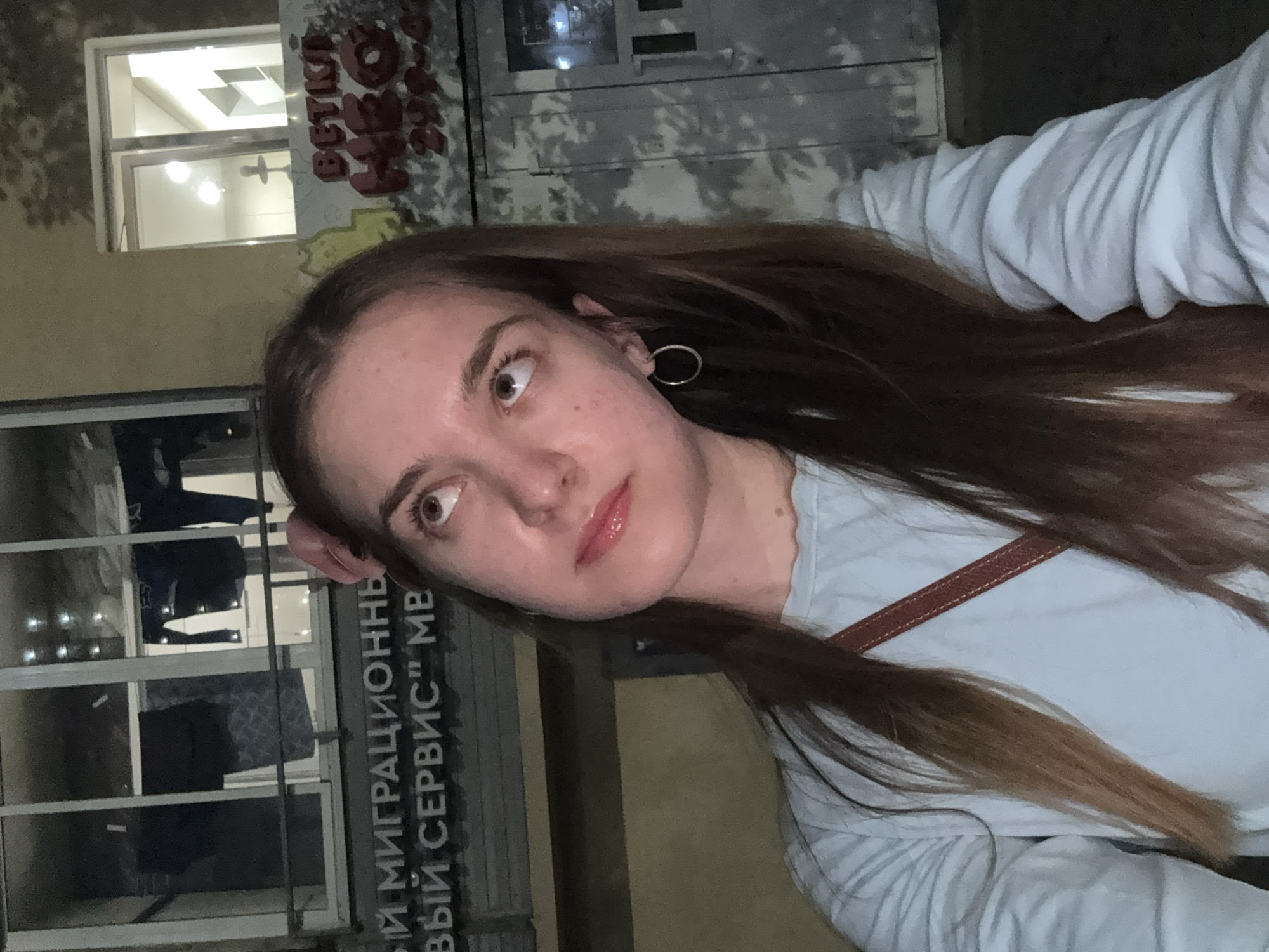 A young woman with long brown hair and fair skin poses for a photo outdoors at night. She wears a light blue shirt and a hoop earring. Behind her is a building with a window display of clothing and a sign in Russian that includes the words "Миграционный Сервис" (Migration Service). Graffiti is visible on the wall above the window. The location is likely in Perm, Russia, given the Russian signage and the woman's appearance.
