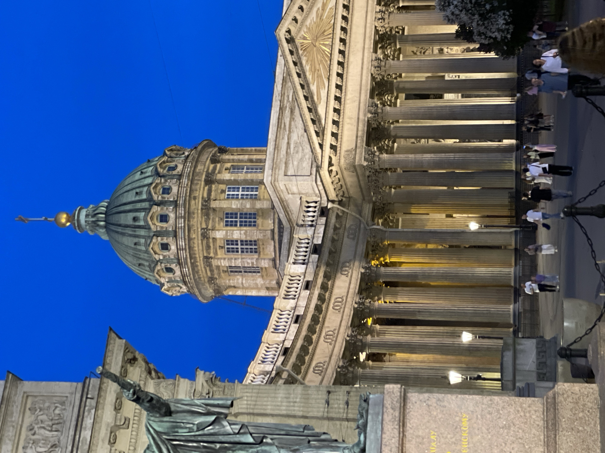 This image captures the Kazan Cathedral in Saint Petersburg, Russia, at dusk or night. The prominent architectural structure, centrally positioned, features a large green dome topped with a golden cupola and a cross, illuminated by external lighting. Below the dome, a circular drum with windows transitions into the main facade, characterized by an extensive semi-circular colonnade with numerous classical columns. A triangular pediment adorned with a radiant eye symbol is visible on the right side of the main building, also illuminated.
In the left foreground, a bronze statue of Field Marshal Mikhail Kutuzov, identified by partially visible Cyrillic text "КУТУЗОВУ" on its stone pedestal, stands partially obscured. The statue depicts a robed figure with its right arm extended upwards. The pedestal itself is a light-colored stone with carved decorative elements, including a low-relief frieze at its upper section and a classical pattern at its base.
The sky above is a uniform deep blue, indicative of twilight or nighttime conditions. The architectural elements of the cathedral are highlighted by warm, artificial lighting. Multiple street lamps with glowing warm lights are distributed across the paved foreground area.
A pedestrian walkway or square occupies the lower portion of the image. Approximately two dozen individuals are scattered across this area, walking in various directions. They are casually dressed in contemporary clothing, appearing to be adults and a few children. No distinct interactions or specific actions beyond casual strolling are discernible. Dark metal chains connect dark bollards or planters along the edge of the pedestrian area in the immediate foreground. On the far right, a dark green shrub or tree with some light-colored foliage is present. The ground is paved with a dark surface.