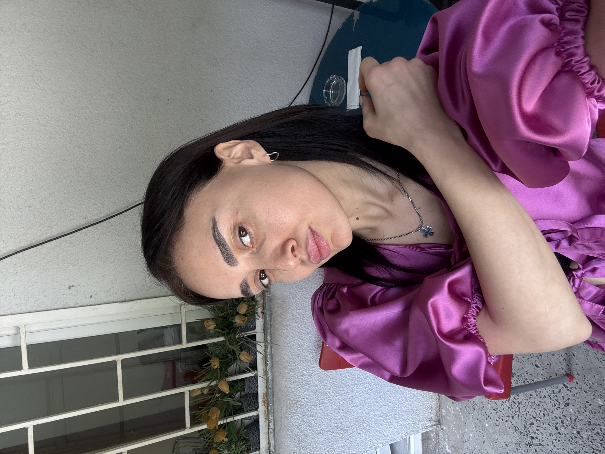 A woman with dark hair, light skin, and strong eyebrows is seated in front of a window with metal bars. A floral arrangement is present behind the bars. She is wearing a purple satin blouse. She has a silver necklace with a pendant. A red chair is partially visible. The background includes a blue object and a light-colored wall. Based on the architectural features and window style, it is possible this photo was taken in Tbilisi, Georgia.