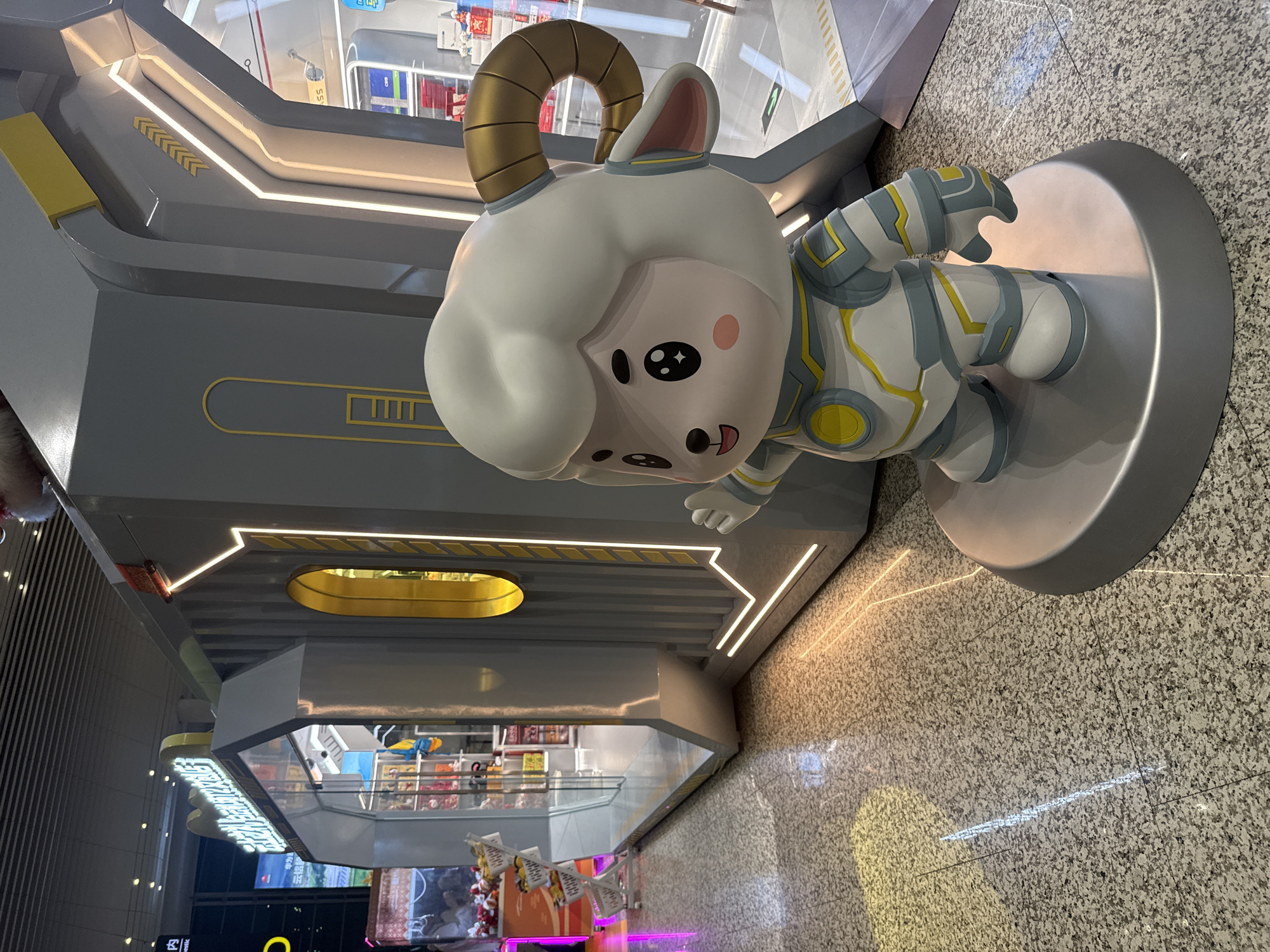 A low-angle, full-color photo depicts a cartoon sheep figure wearing an astronaut suit, situated in a shopping mall in Guangzhou City, China. The sheep is mounted on a gray, circular platform that rests on a light brown, speckled tile floor. The background features the interior of the mall, including a retail store with displays visible behind a glass barrier. The walls and architectural elements surrounding the figure are painted in shades of gray, accented with yellow geometric patterns and illuminated with strips of linear lighting. The lighting casts reflections onto the tiled floor, adding depth to the scene. The overall scene appears to be well-lit and modern in its design.
