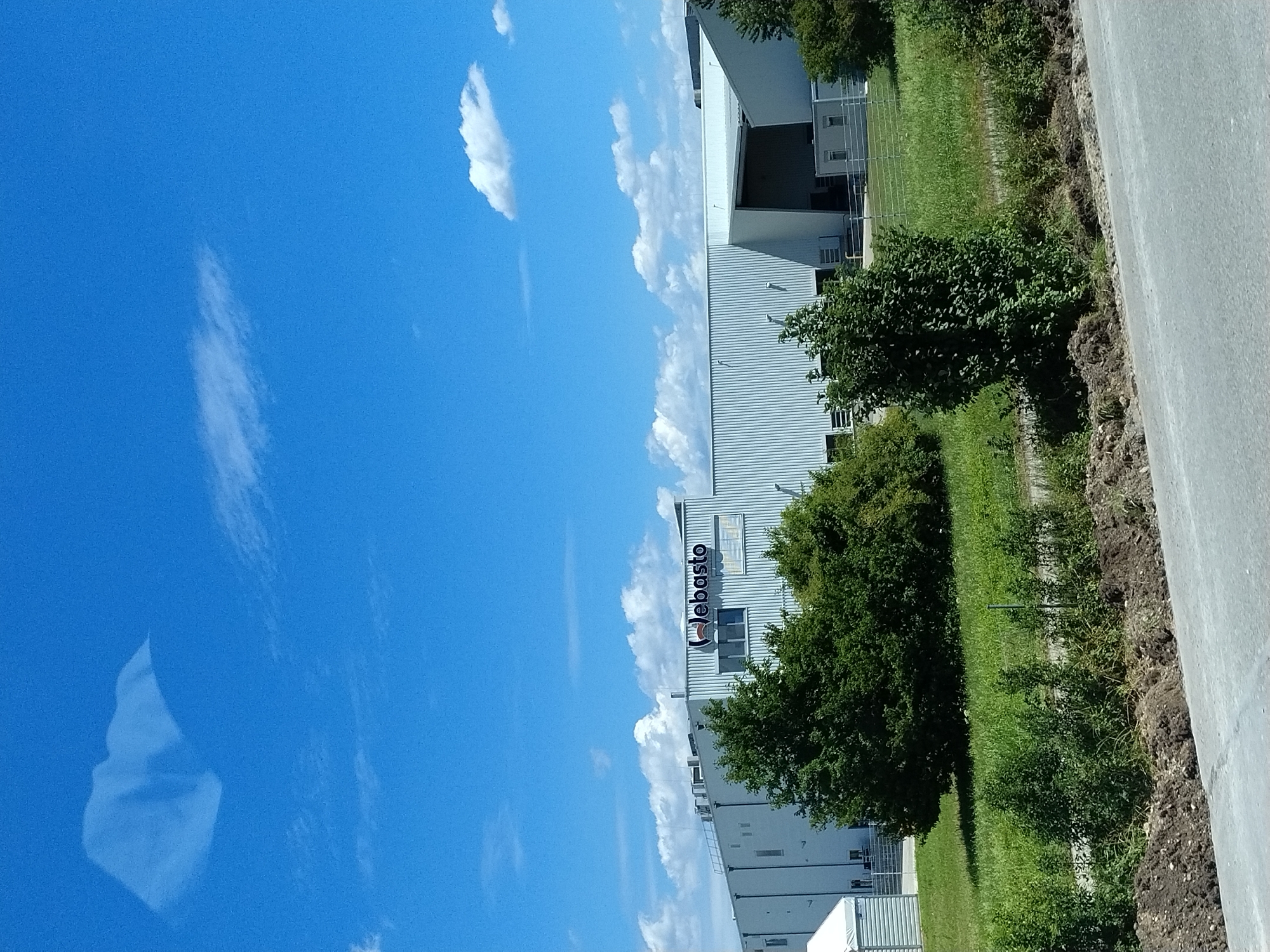 The image shows a large, modern industrial building with the "Webasto" logo visible on its facade. The building is white and appears to be constructed with corrugated metal siding. In front of the building, there is a patch of green grass with several trees. The sky is blue with scattered white clouds. The scene is likely an exterior shot of a Webasto facility in Hengersberg, Germany, based on the provided context. There are no people or specific actions depicted in the image. The environment suggests a sunny day.