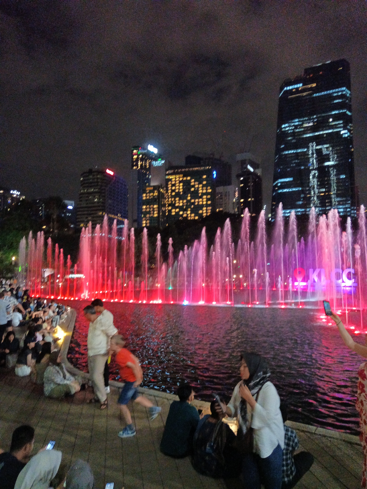 This image depicts a nighttime scene at the KLCC Park in Kuala Lumpur, Malaysia, a popular urban oasis adjacent to the Petronas Twin Towers. In the foreground, a group of people are gathered on a wooden boardwalk surrounding a large water fountain. The fountain features numerous spouts of water illuminated by vibrant pink and red lights, creating a dynamic display against the dark sky.
Several individuals are visible, including a family with a child running near the fountain. One person in the foreground is holding a mobile phone, seemingly taking a photograph or video of the fountain show. Another woman, wearing a hijab and a white top, is also holding a phone. In the background, the iconic Petronas Twin Towers and other city skyscrapers are illuminated, showcasing Kuala Lumpur's impressive skyline. The overall atmosphere is one of a lively public gathering enjoying the evening entertainment.