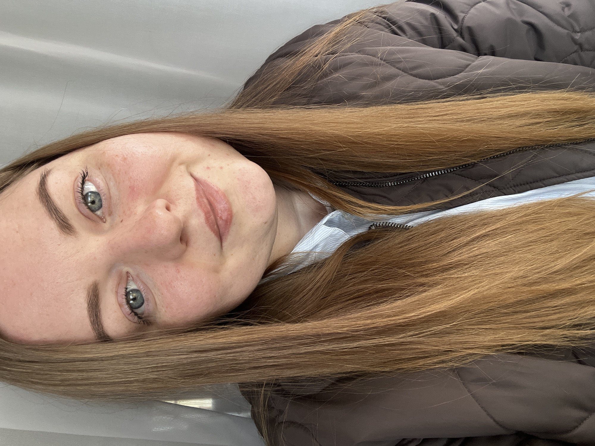 A close-up portrait shows a young woman, oriented with her head on the left side of the frame, looking directly towards the viewer with a gentle, subtle smile. Her skin is fair, featuring freckles across her nose and cheeks, and some natural redness. She has striking blue-grey eyes, enhanced with mascara on her eyelashes, and dark, well-defined eyebrows. Her lips have a light, natural-toned tint.
Her long, straight hair, which is a light brown or golden blonde color, cascades over her shoulder and chest. She is dressed in a dark brown, quilted jacket with a visible zipper detail down the front. A light-colored, possibly striped, collared shirt is visible underneath the jacket. The background is plain and light, suggesting an indoor setting. There is no visible text in the image. This media file is from Раздолье, Russia.