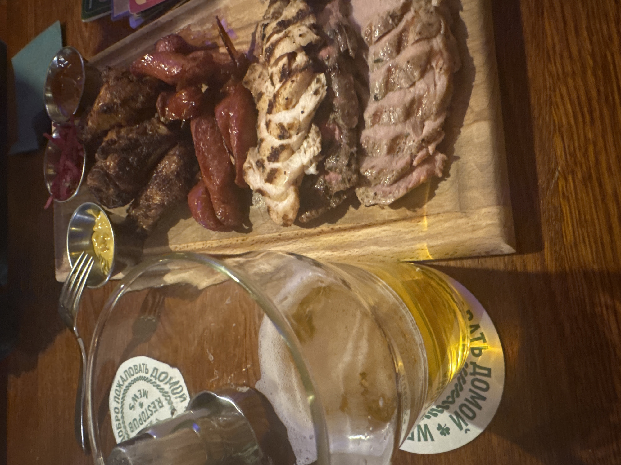 A high-angle shot shows a wooden table. At the top of the frame, a rectangular wooden board holds an assortment of cooked meats including sausages, grilled chicken, and sliced pork. There are three small metal dishes containing condiments; mustard, a red sauce and another containing a red pickled ingredient. In the lower portion of the image, a partially consumed glass of beer sits on a coaster with the words "ДОБРО ПОЖАЛОВАТЬ ДОМОЙ RESTOPUB NEW" indicating the scene is likely at a restaurant/pub named "New Restopub" which can be loosely translated from the Russian as "Welcome Home." The city of Saint Petersburg is inferred because the Russian text on the coaster is written in the language and font style commonly associated with the location. A fork rests near the beer glass.