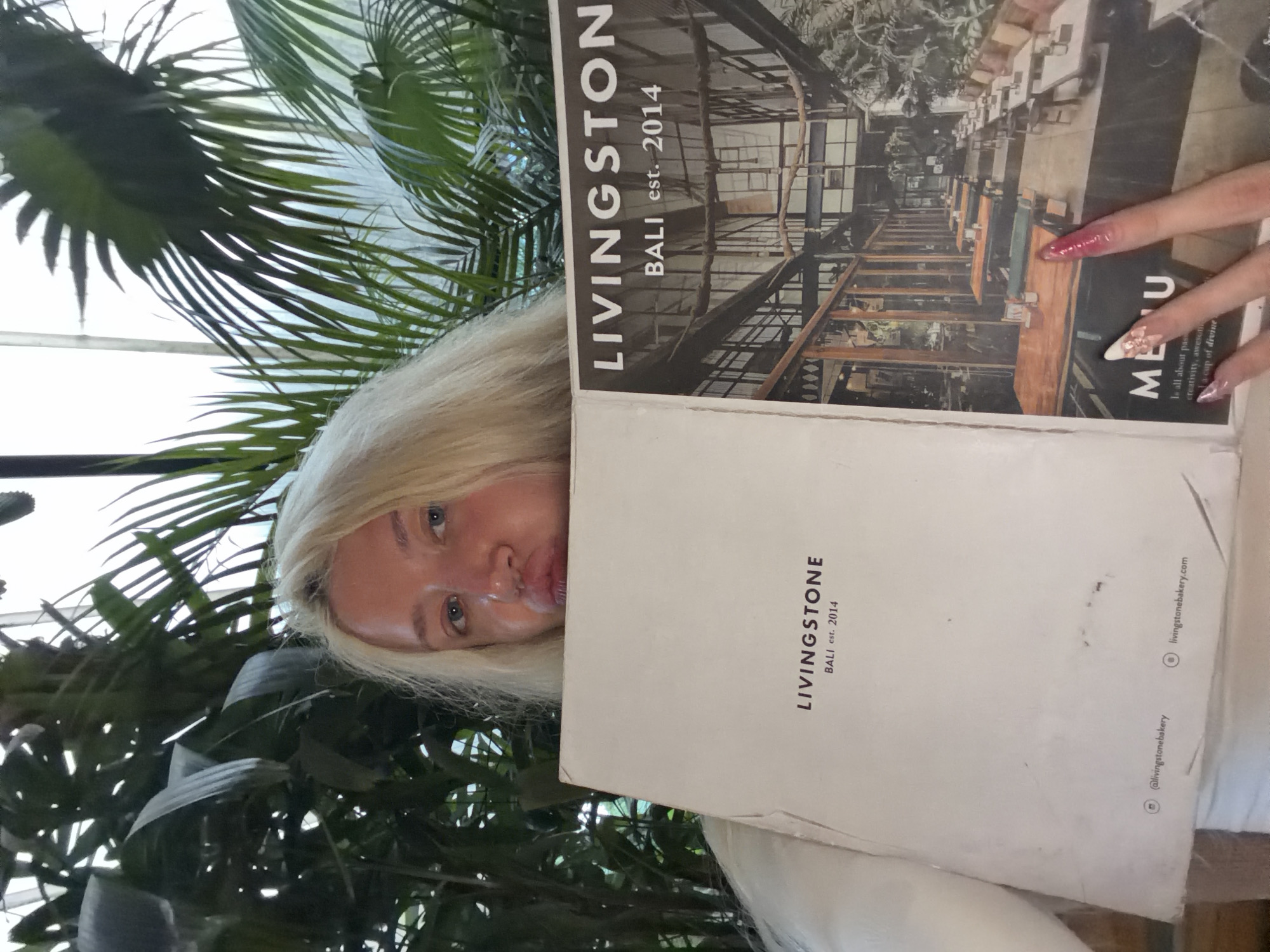 In North Kuta, Indonesia, a blonde woman is posing with a menu from Livingstone, a establishment established in 2014. She is partially obscured by a large menu, which features an image of the interior of the restaurant with tables and chairs. The menu also displays the text "LIVINGSTONE BALI est. 2014" and website information. Lush green plants surround the woman, suggesting an outdoor or garden-like setting.
