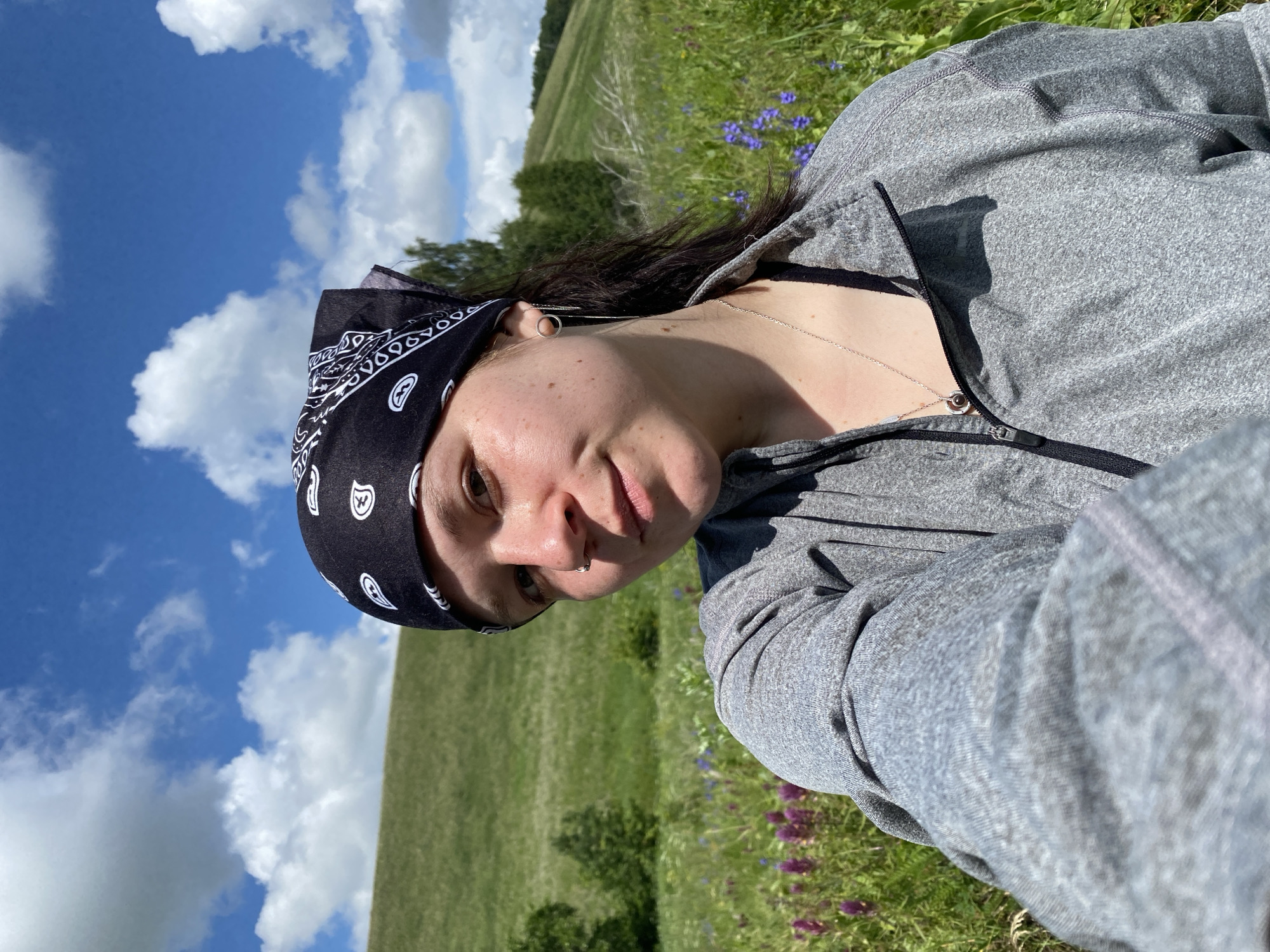 A young person with a black bandana featuring a white paisley pattern is taking a selfie in a field of green grass and purple and blue wildflowers under a cloudy blue sky. The person is wearing a grey zip-up jacket over a black tank top and a silver necklace with a dark pendant. They have a nose ring and a long, delicate earring. The background shows a gently sloping green hill. The image appears to be taken during the daytime. The location is identified as the city of Bogoslovka, Russia.