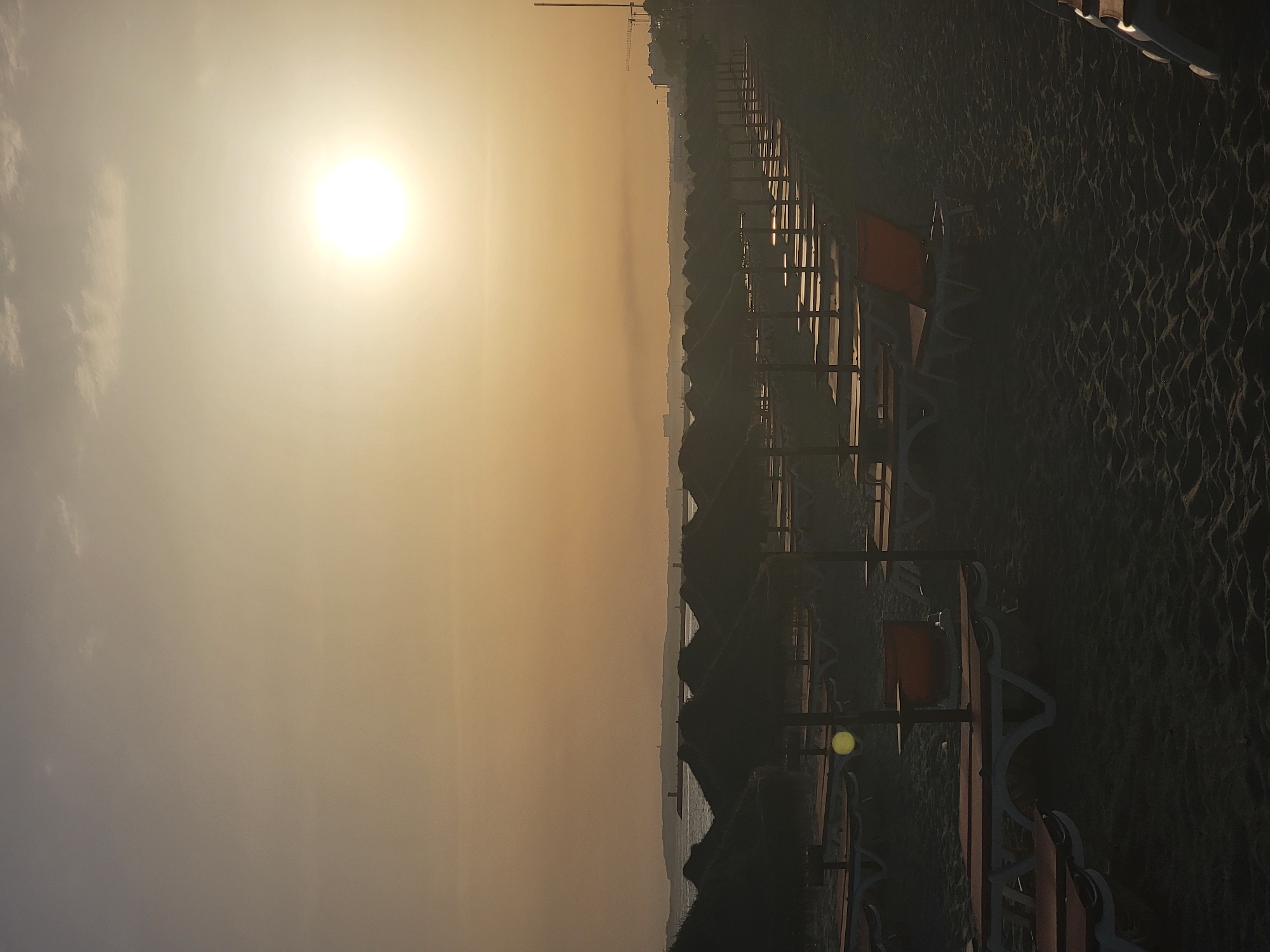 This is a full, vertical shot of a beach in Quarteira, Portugal at either sunrise or sunset. The sun is a bright, hazy orb in the upper right quadrant of the image, casting a warm, orange glow across the sky and a muted light on the scene. In the foreground, rows of empty, dark beach loungers stretch into the distance. Each lounger has an orange cushioned back. Behind the loungers are rows of thatched umbrellas, also empty. The sand appears dark and textured. In the mid-ground, a hazy body of water can be seen, with a distant landmass on the horizon. Structures, possibly buildings and a lighthouse, are faintly visible on the horizon line. The overall atmosphere is tranquil and deserted, suggesting the early morning or late evening.