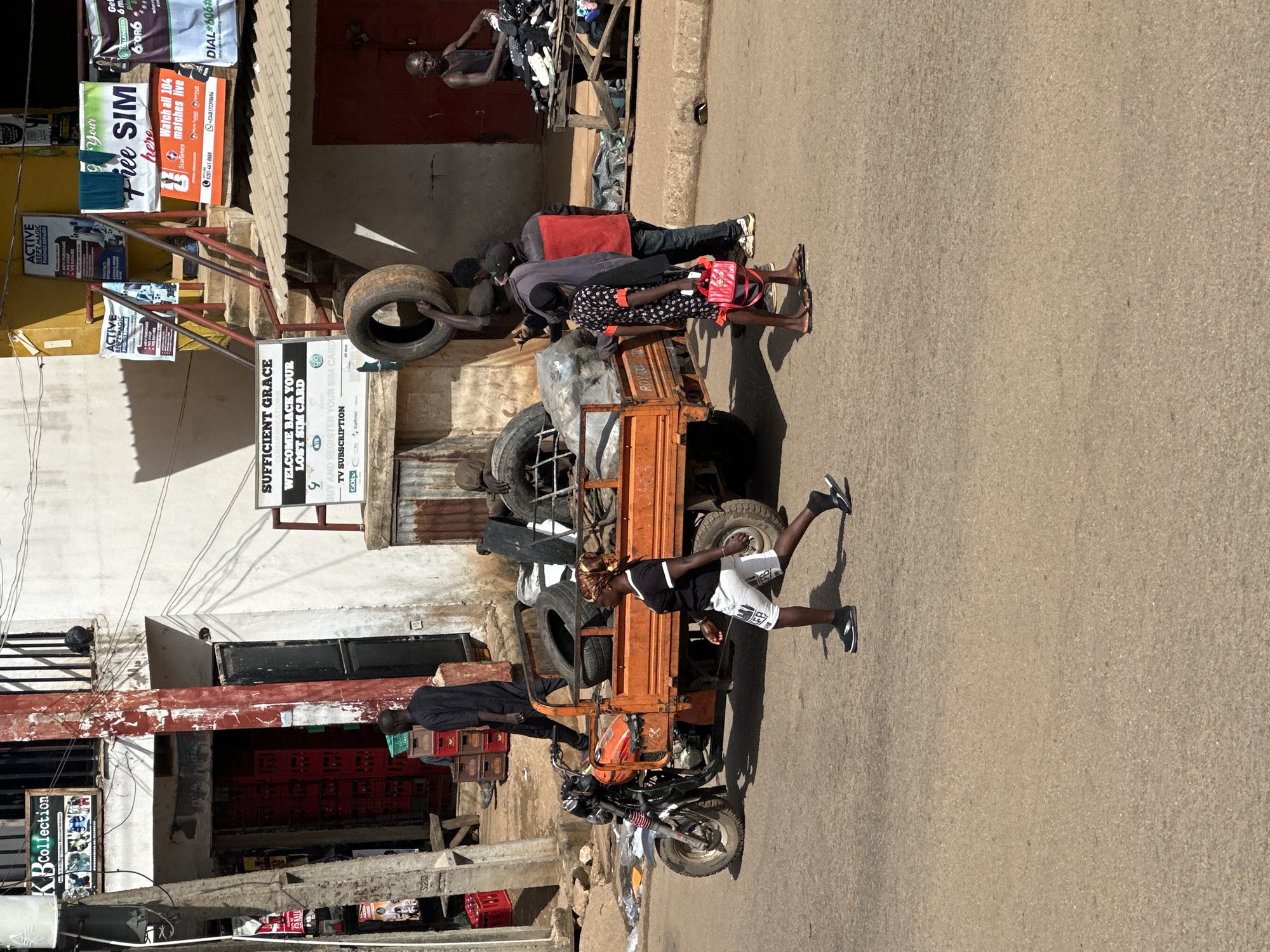 A sunny, outdoor scene on a paved road in Bukuru, Nigeria depicts a street market. In the foreground, a woman in white shorts and a black t-shirt walks across the road. Behind her, an orange three-wheeled truck is parked, loaded with tires. Several people are gathered around the truck and a nearby stall. To the left, a building with red crates stacked outside a doorway is visible. To the right, a man stands behind a table piled with shoes. Signs advertise various services, including phone repairs and TV subscriptions. The overall impression is one of a busy commercial area.