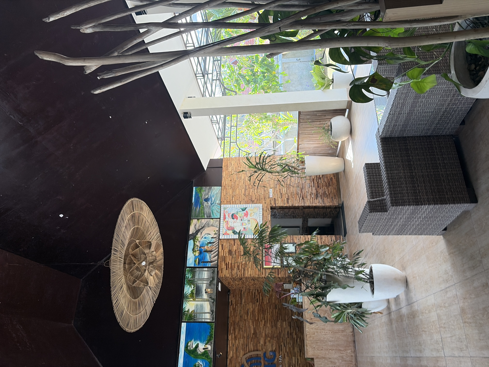 The image depicts the interior of a resort lobby in Nusa Penida, Indonesia. The lobby features a high, dark brown ceiling with a large, woven bamboo pendant light fixture. A reception desk with a brick facade is visible on the left, adorned with several framed paintings and a large logo. The floor is tiled with light-colored square tiles. To the right, there are two woven rattan armchairs and a glass-topped coffee table, suggesting a seating area. Numerous potted plants, including large leafy ones in white planters, are strategically placed throughout the space. In the background, large windows reveal lush green vegetation and the bright tropical sunlight. The overall atmosphere appears to be relaxed and inviting, typical of a tropical resort.