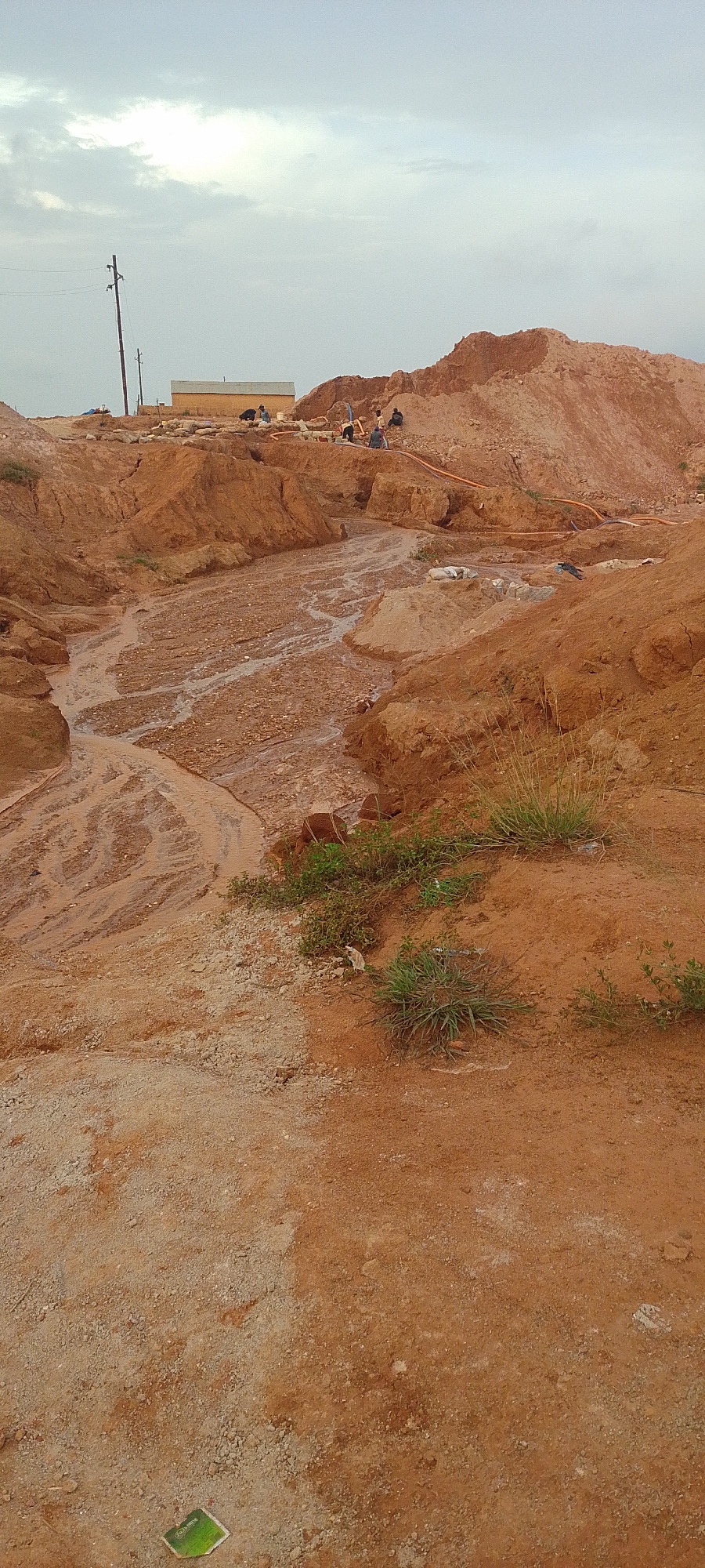The image depicts an outdoor construction or excavation site under a cloudy sky. A muddy, channelized stream flows through reddish-brown earth and gravel. Several individuals are visible working on a mound of earth in the background, some appearing to be using tools and possibly operating hoses. A light-colored, rectangular structure is situated behind them, and a utility pole with electrical wires stands to the left of the frame. The foreground consists of a barren, reddish-brown soil surface with scattered gravel. In the lower left corner, a small, green object, possibly a piece of plastic or packaging, is visible. The overall impression is of an active site with earthmoving and potential water management activities.