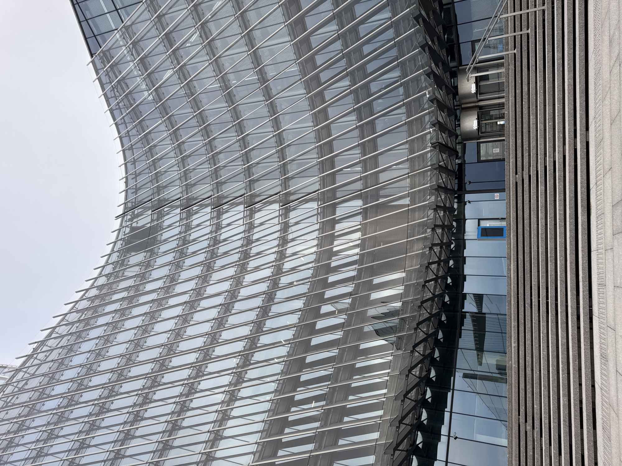 This is an exterior, eye-level, vertical, medium shot of a modern architectural building. The building has a curved facade made of glass panels and metal beams, reflecting the overcast sky. To the right of the curved glass facade is a section of the building with a more rigid, vertical design, featuring a darker, textured material and vertical grooving. Between the curved glass section and the vertical section, there are visible windows and what appears to be an elevator shaft with metallic railings. One of the windows on the elevator shaft has a bright blue rectangular frame. The scene is dominated by cool tones of grey and blue, suggesting a daytime shot on a cloudy or hazy day. There are no people visible in the image, only the imposing structure of the building. The overall impression is one of sleek, contemporary design. There is no visible text.