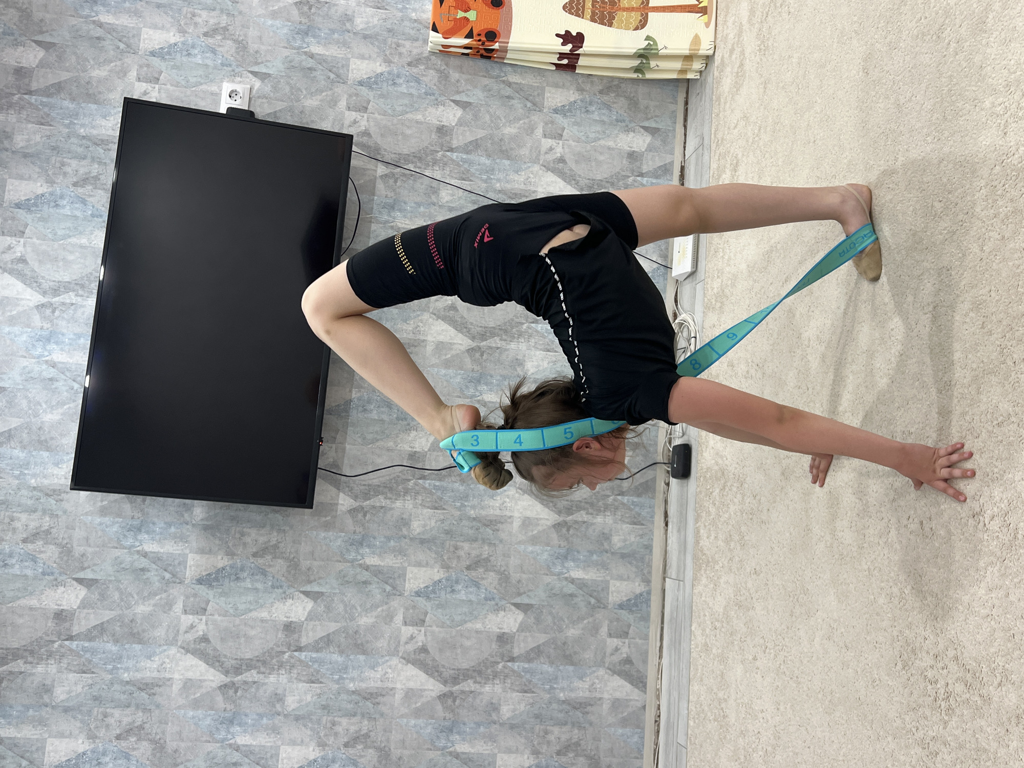 A young female is in a backbend pose in a room. She is wearing black shorts, a black shirt, and ballet shoes with a blue stretching strap around her head and left foot. She is facing away from a flat screen television mounted on a wall with patterned wallpaper. The floor is covered with a tan, textured carpet, and there is a folding mat visible in the background. The location is indoors, likely a home, but the city of Chaikovsky, Russia cannot be determined from the visual information.