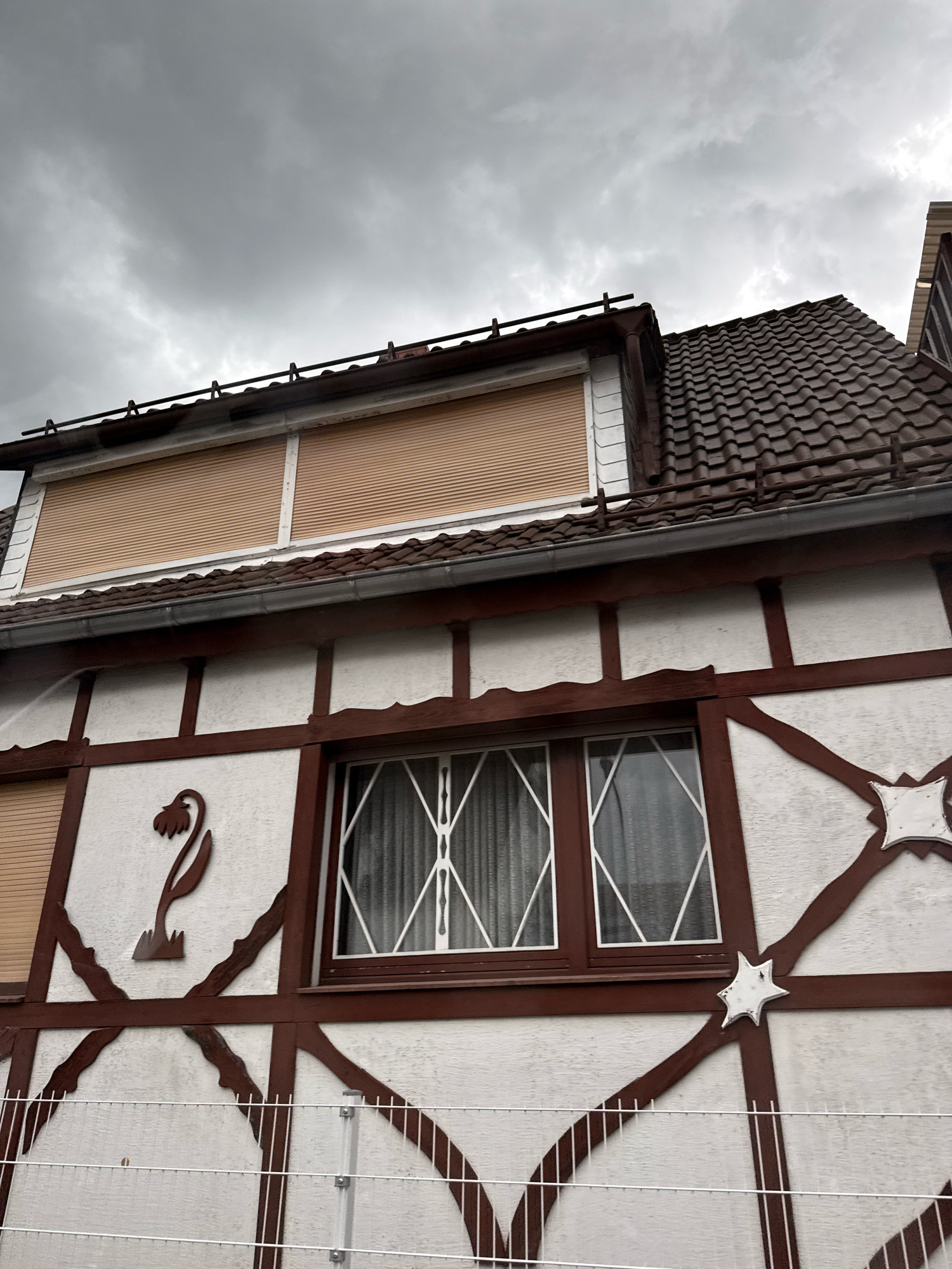 This is a low-angle, close-up shot of the facade of a building with a traditional timber-frame design, likely located in Bad Sachsa, Germany, under a cloudy, overcast sky. The building features a white stucco exterior with dark brown timber beams forming decorative patterns, including an abstract flower motif and two star-shaped ornaments. The upper part of the building shows a roof with dark, wavy tiles and a section with what appear to be closed beige roller shutters. A window with diamond-shaped decorative white bars and sheer white curtains is visible on the lower floor. In the foreground, a white metal fence with horizontal wires is present. The overall scene suggests a slightly damp or gloomy weather condition.