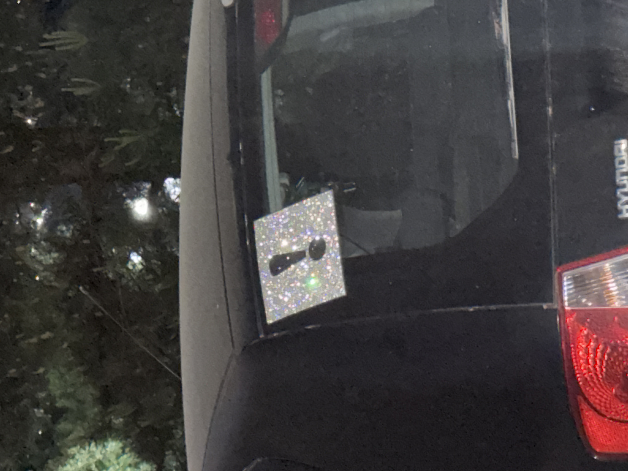 The image shows the rear of a dark-colored Hyundai car, specifically the rear window and taillight area. A square, glittery sticker featuring a silhouette of a spoon is adhered to the rear window. The location is suggested to be Izhevsk, Russia (based on the model of car). The car is parked in an outdoor environment, with a dark background suggestive of nighttime and foliage visible out of focus on the left side. The taillight is partially visible on the right side of the frame.