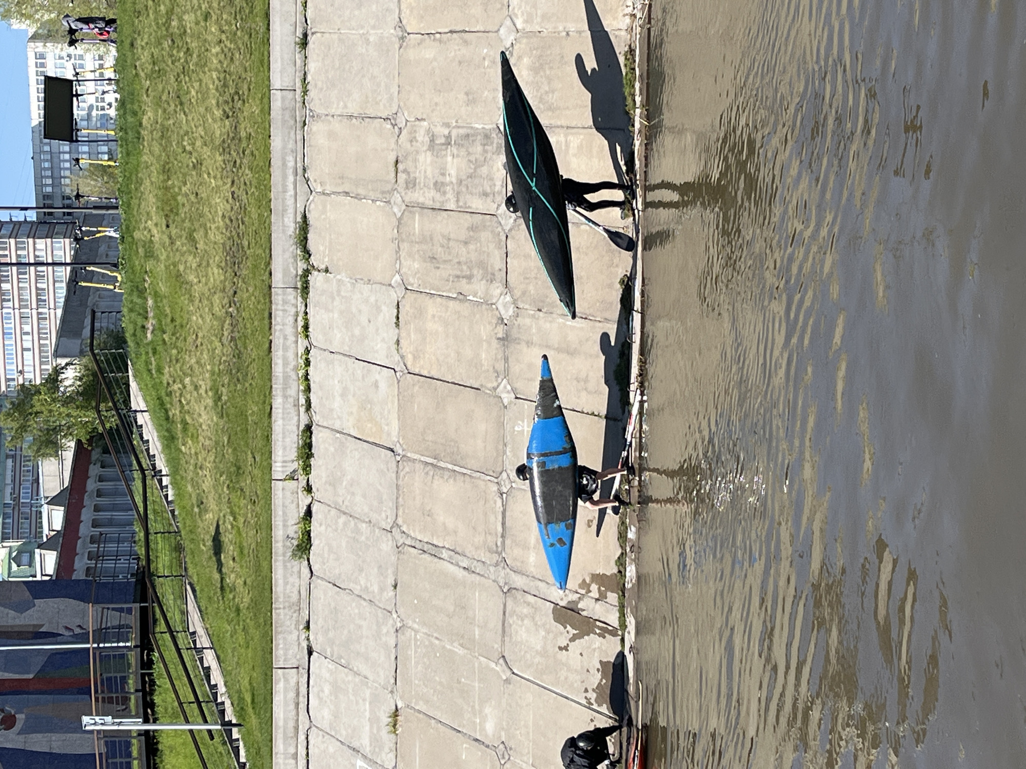 This aerial view shows two individuals preparing to kayak on the Yenisey River in Krasnoyarsk, Russia. Two kayaks are visible on the concrete embankment; one is black with a neon green design, while the other is blue with black. Shadows suggest the individuals are positioned near the kayaks, ready to enter the water. The river water reflects the sunlight. Adjacent to the embankment is a grassy area and a staircase with a black railing. In the background are buildings and trees. A few people are visible further away.