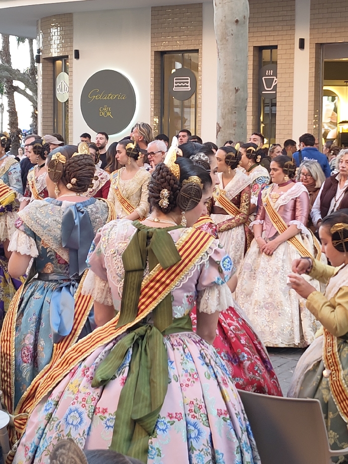 The image captures an outdoor gathering in Dénia, Spain, specifically in front of a "Gelateria Carte D'Or" establishment. The foreground and middle ground are populated by approximately ten women and one young girl, all dressed in elaborate traditional Valencian costumes. These costumes feature voluminous, intricately patterned brocade and floral dresses in shades of pink, light blue, cream, and maroon. Each individual wears a red and yellow striped sash diagonally across the chest and exhibits an elaborate traditional hairstyle with multiple braided buns adorned with decorative combs.
In the immediate foreground, facing away from the viewer, are two women: one in a pink floral dress secured with a dark green bow at the back, and another in a light blue floral dress with a corresponding light blue bow. A young girl, partially visible on the right, wears a yellow top and holds a small white object. The background reveals a mixed crowd of onlookers in contemporary clothing. The setting includes a modern commercial building with a textured light-colored facade and large glass windows. A prominent circular sign displays "Gelateria Carte D'Or" in gold lettering, flanked by icons representing an ice cream cone, a cake, and a steaming cup. A large tree trunk stands in the center-right of the frame, and a portion of a dark chair is visible in the bottom right. The scene is illuminated by daylight.