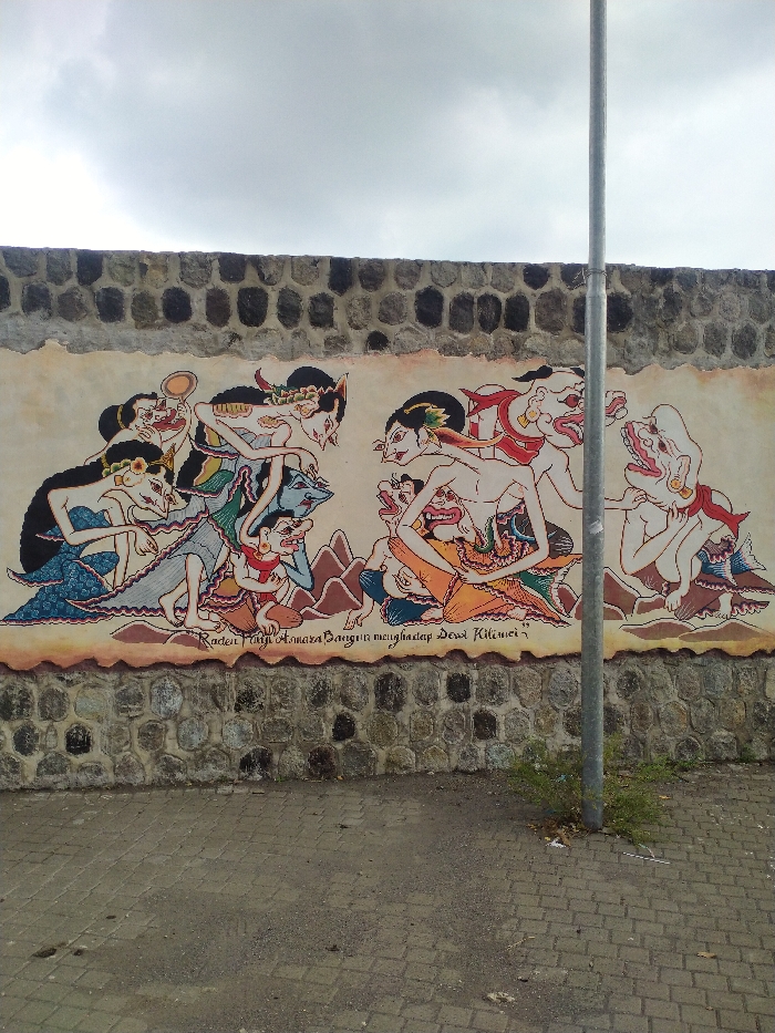 A vibrant mural, painted in the traditional Javanese wayang style, adorns a stone wall in Karanganyar, Indonesia. The artwork depicts a dynamic narrative scene featuring numerous stylized figures. On the left, several characters, some with intricate headdresses and delicate features, are engaged in various poses, including one holding a round object resembling a mirror. The central part of the mural shows a key interaction between figures, one of whom is identified by the accompanying text. To the right, more grotesque or demonic-looking figures with large, open mouths and exaggerated features are visible.
Below the central figures, text written in white states: "Raden Panji Amara Bangun menghadap Dewi Kilisuci." This translates to "Raden Panji Amara Bangun meets Dewi Kilisuci," indicating a specific mythological or historical encounter. The figures are rendered with distinctive colors, including blues, reds, golds, and greens, and wear traditional clothing and ornaments.
The mural is painted directly onto a section of a robust stone wall, constructed from irregularly shaped dark grey and brown rocks. Below the wall, the ground is paved with grey interlocking bricks, suggesting a sidewalk or public pathway. A tall, slender metal pole, possibly a street light post, stands on the right side of the frame, partially obscuring the mural. The sky above is overcast with a blanket of grey clouds, indicating a cloudy daytime.