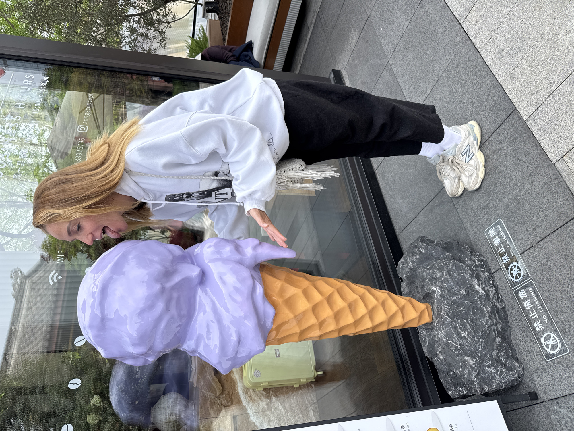 A woman with long blonde hair is standing on a gray tiled sidewalk in front of a storefront with a large glass window. She is wearing a white sweatshirt with black print, black pants, white socks, and white sneakers with a blue "N" on them. The woman is leaning towards a large, sculpted, lavender-colored ice cream cone that is inside the storefront, sticking her tongue out as if to lick it. Her hand is extended towards the ice cream cone.
Inside the storefront behind the ice cream cone, there's a green rectangular object. A large, gray rock sits next to the ice cream cone, partially obscuring the view.
On the sidewalk, there are signs with Chinese characters and English translations that read "NO LEANING" and "NO CLIMBING." There is text in the window behind the woman, including "SS HOURS," "Mon 10:00am-9:30pm," and a phone number, "533926796 songsconcept." The scene appears to be set in Jinjiang District, China.