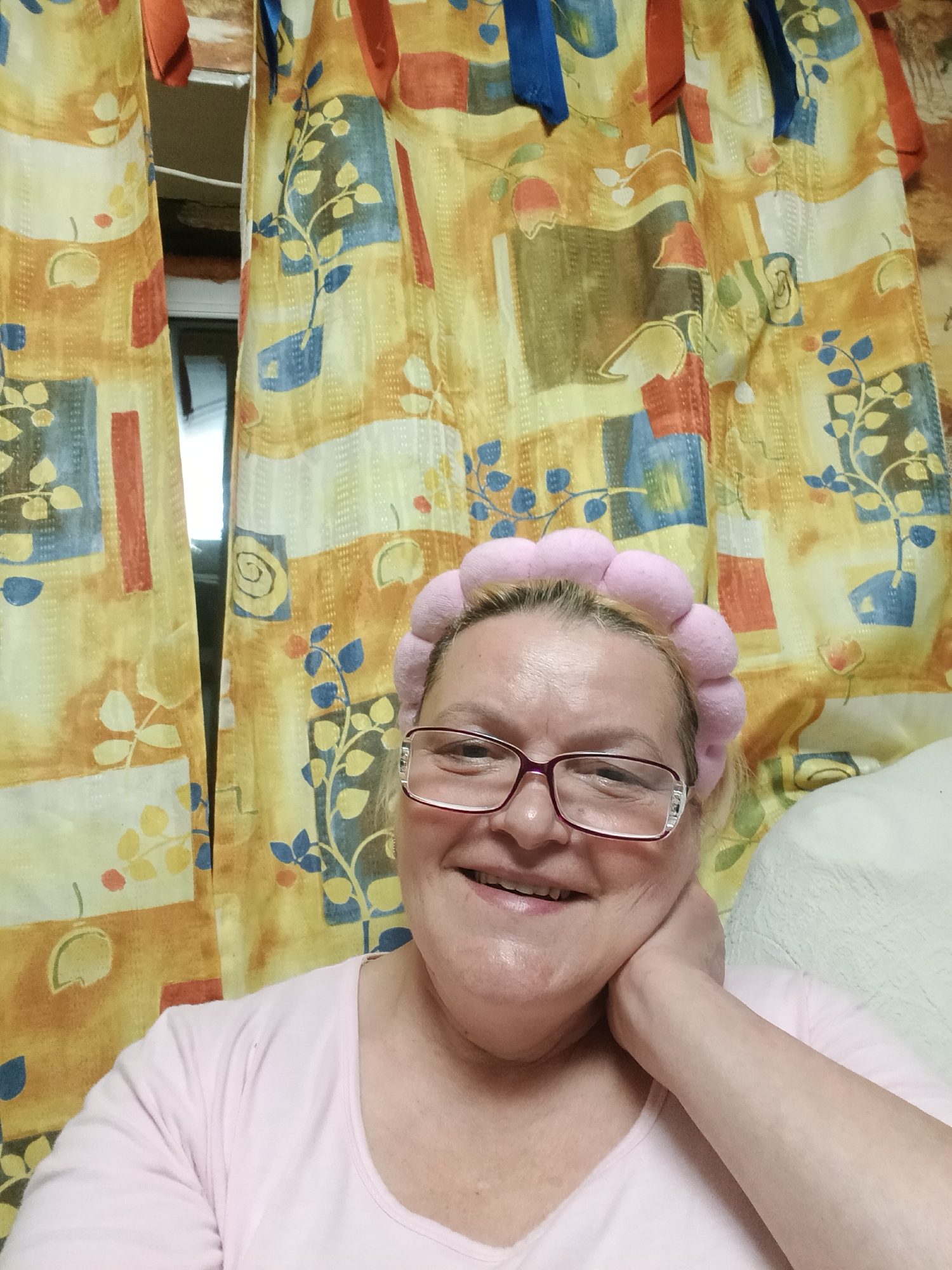 A woman with glasses and a pink headband smiles at the camera. She is wearing a light pink shirt and her right hand is resting on her neck. The background is a patterned yellow and orange curtain. The location is Красный Маяк, Russia.