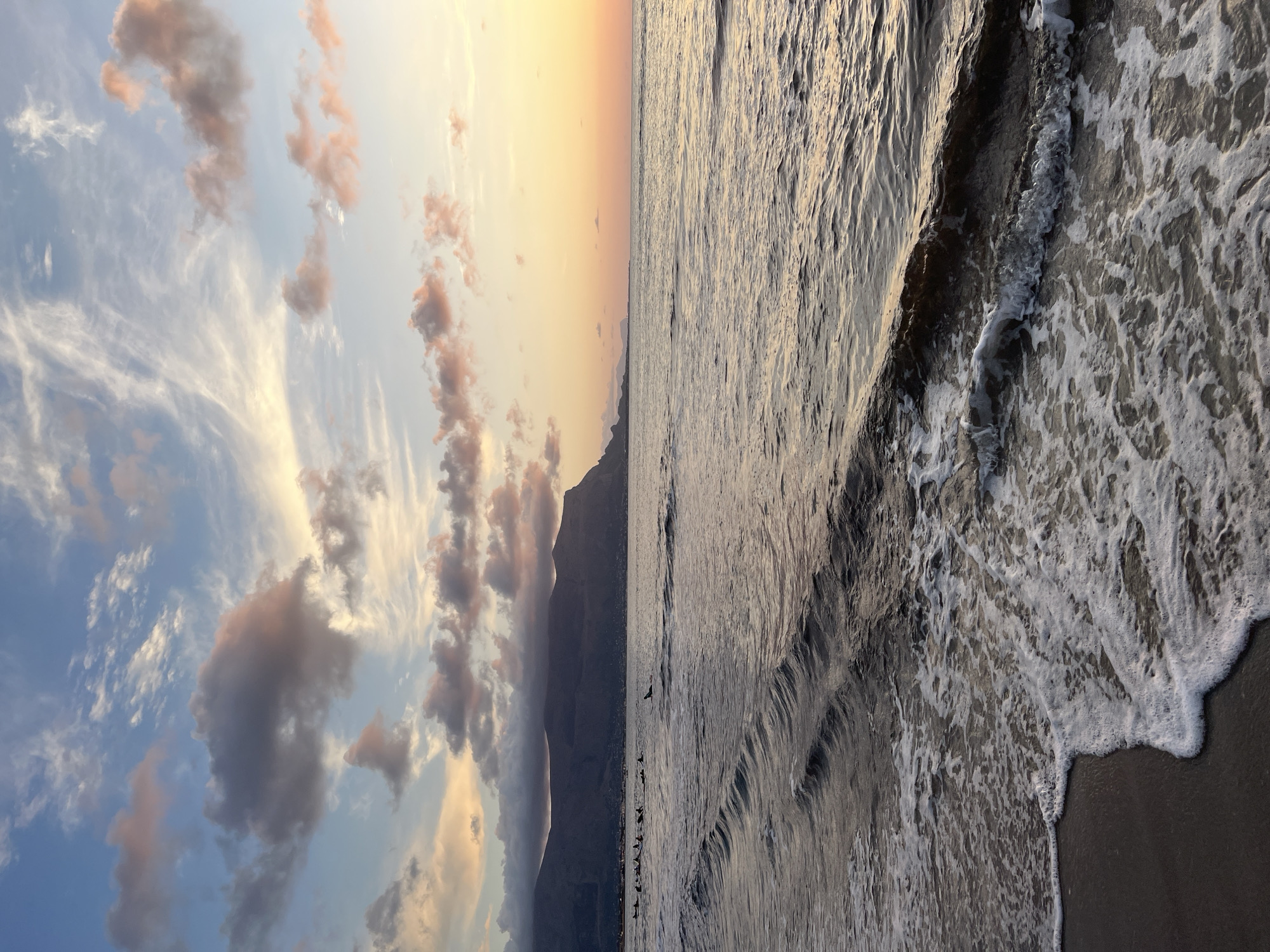 A wide-angle, eye-level shot captures a tranquil sunset over the sea in Isola delle Femmine, Italy. The sky is a canvas of soft pastel colors, with wispy clouds tinged pink and orange by the setting sun, contrasting with patches of blue. In the distance, a range of dark, silhouetted mountains forms the coastline, with a small town nestled at their base. The sea is calm, with gentle waves lapping at the sandy shore, creating patterns of foam. A group of people can be seen in the water, some appearing to be on surfboards, while a single dolphin breaks the surface further out. The overall atmosphere is serene and picturesque.
