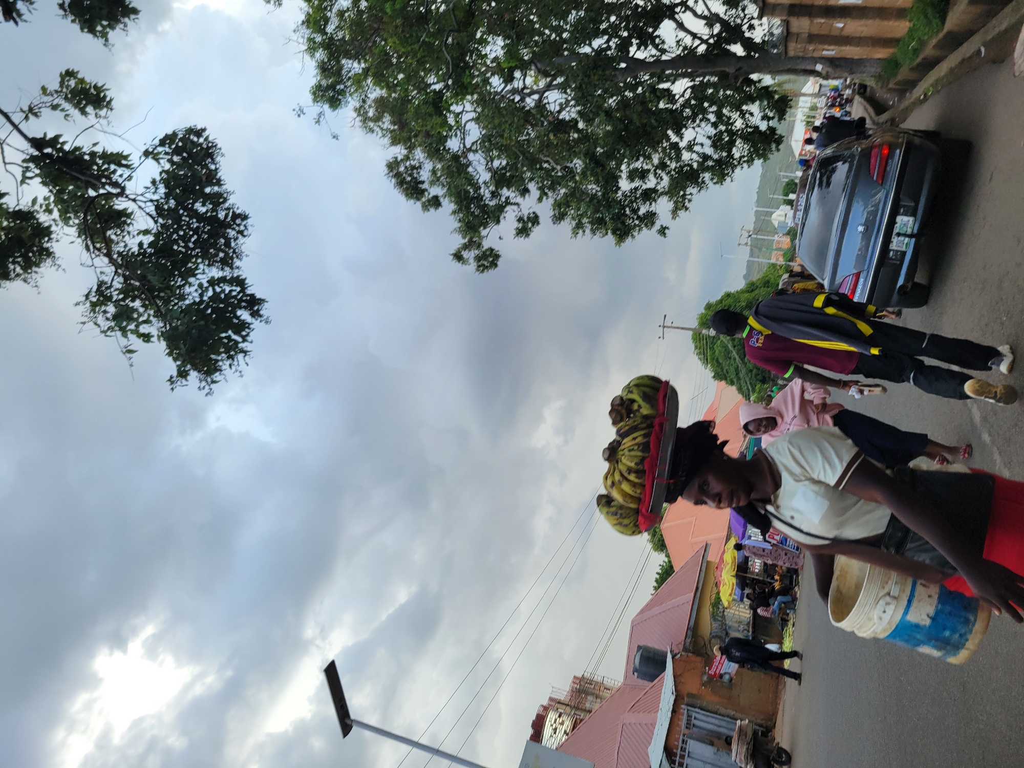 This is a street scene in Jos, Nigeria, featuring a woman carrying a large bunch of bananas on her head, balanced on a tray. She is walking on the right side of the road, carrying a large bucket in her left arm. She is wearing a light-colored t-shirt, a dark skirt, and red shorts underneath. Behind her, another woman in a pink hoodie is walking. In the background, there are several buildings with orange roofs, market stalls with umbrellas, a streetlight, and a dark-colored sedan car driving on the road. A tree with green foliage is visible on the right side of the image, and the sky is overcast with clouds.